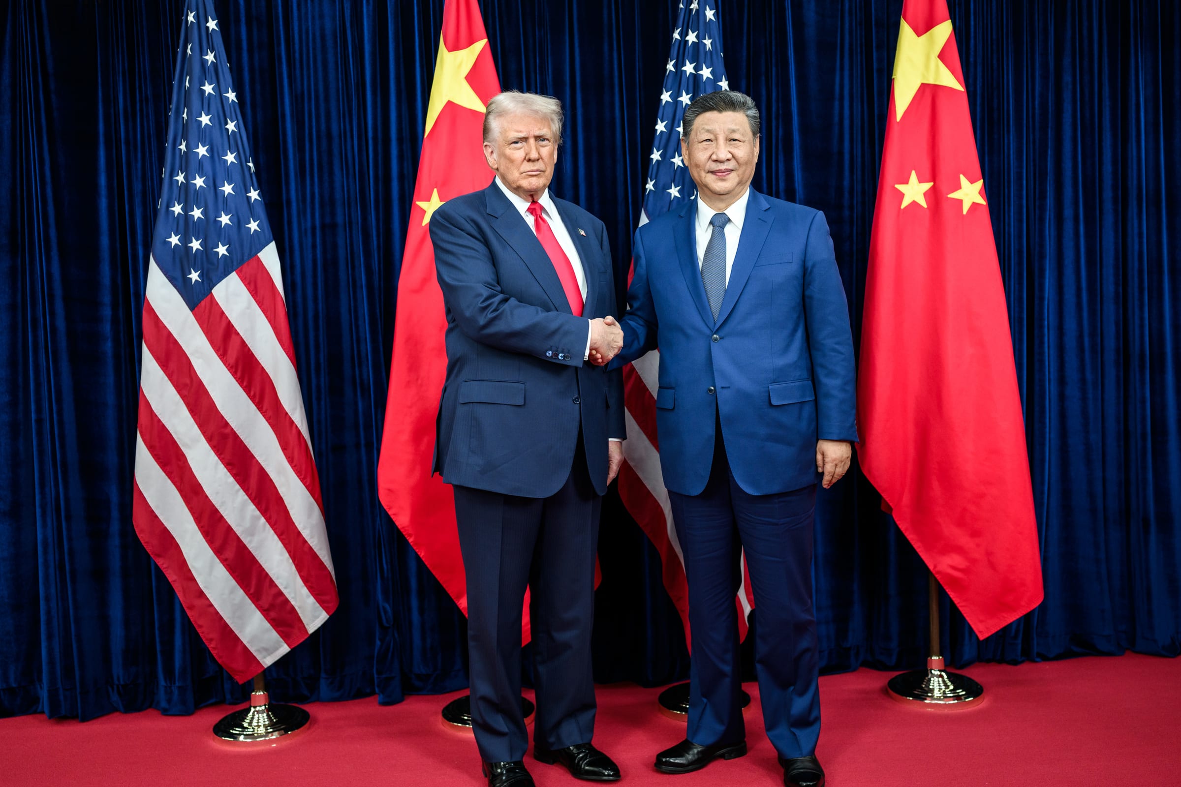 Donald Trump and Xi Jinping shake hands in front of alternating US and Chinese flags at a bilateral meeting.