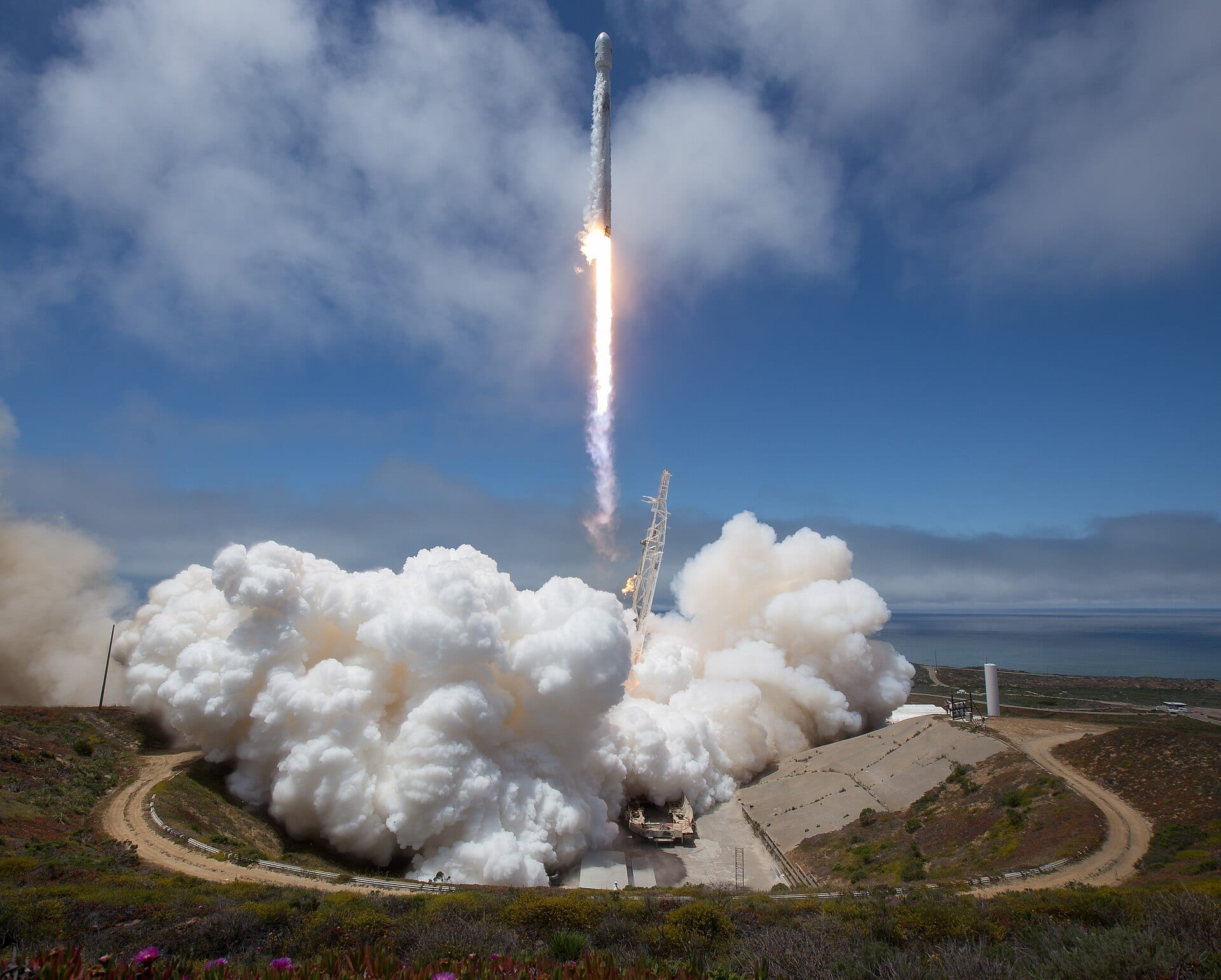 A SpaceX Falcon 9 rocket climbs away from its Vandenberg launch pad, flame trailing above a huge white exhaust cloud.