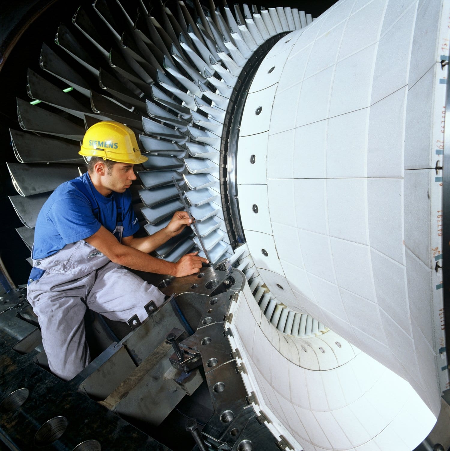 A Siemens technician in a yellow hard hat assembles the blades of a large industrial gas turbine.