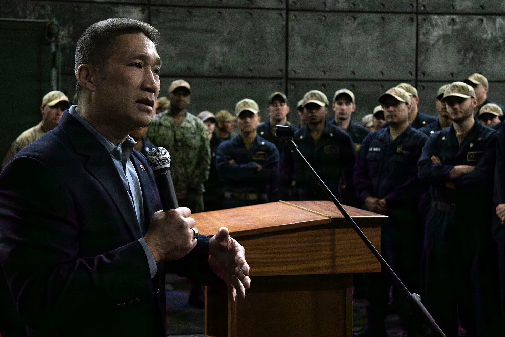 Acting Navy Secretary Hung Cao speaking into a microphone before a formation of sailors aboard a warship