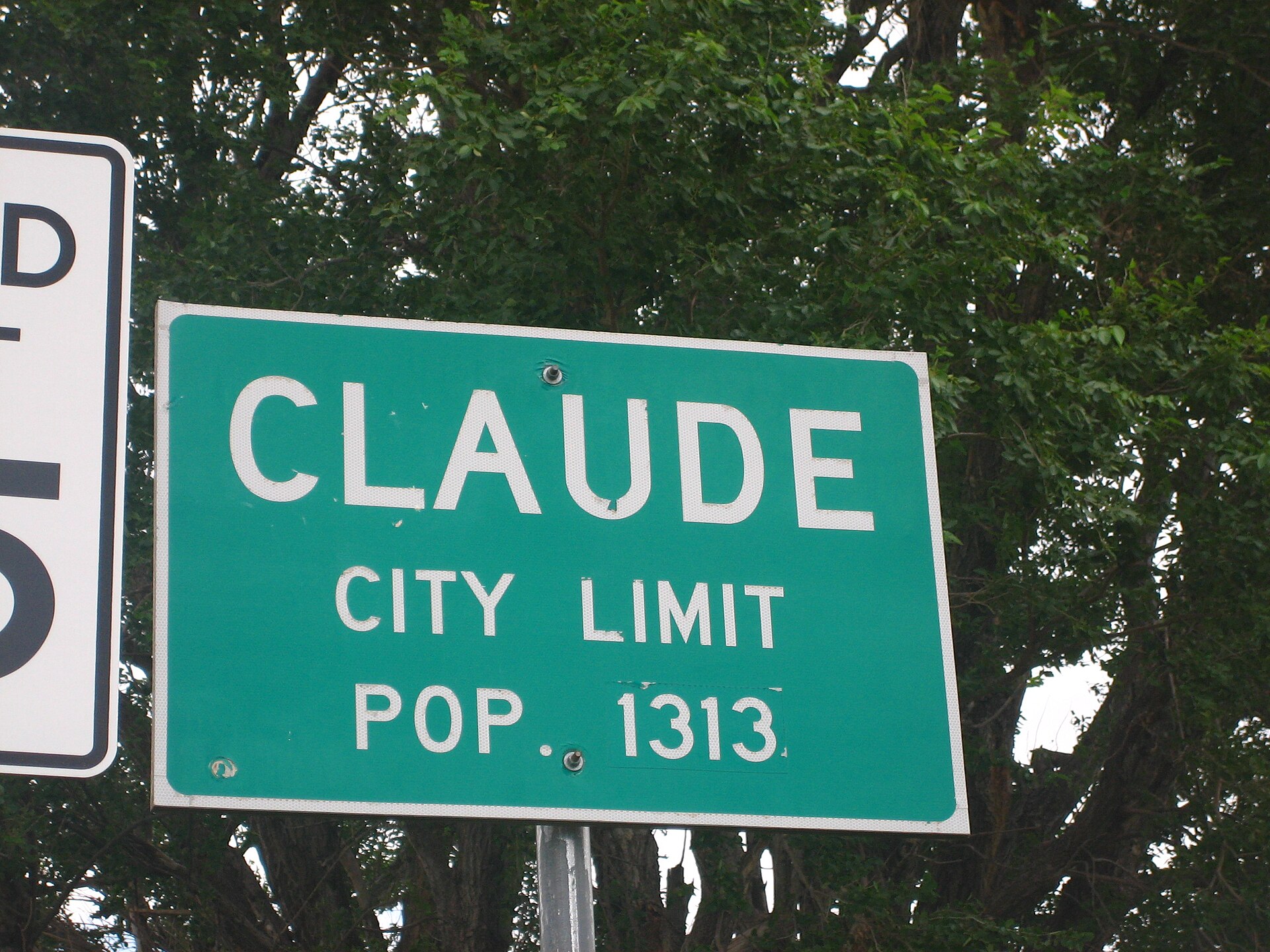 Green highway sign reading Claude, City Limit, Pop. 1313, in front of trees