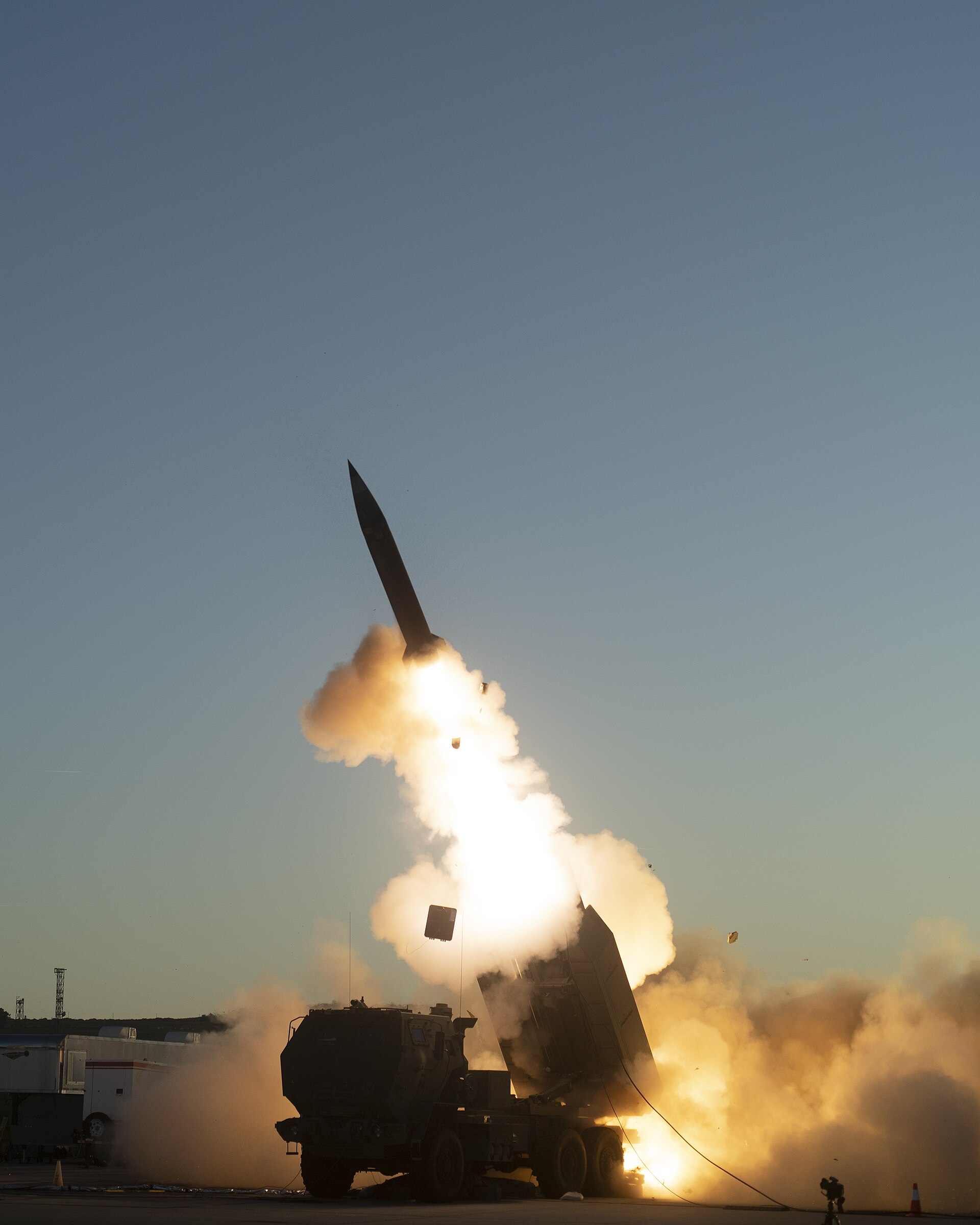 A Precision Strike Missile lifts off from a HIMARS launcher at dusk, flame and smoke trailing beneath it