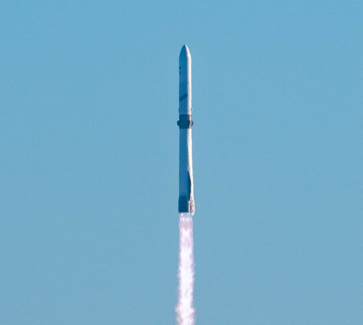A New Glenn rocket climbs into a clear blue sky on a column of exhaust, its black-striped booster visible below the white upper stage.