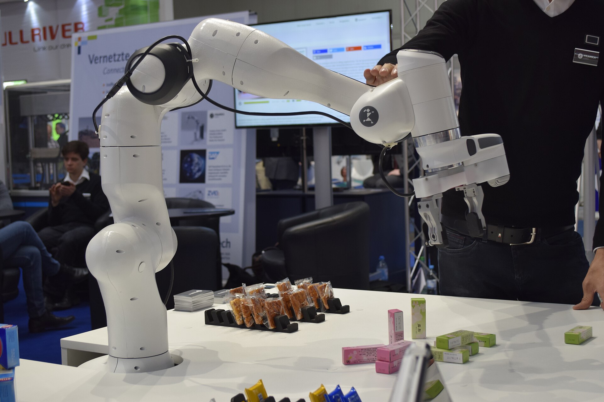 A Franka Emika robot arm in side view at a trade fair booth, reaching over a table of snacks