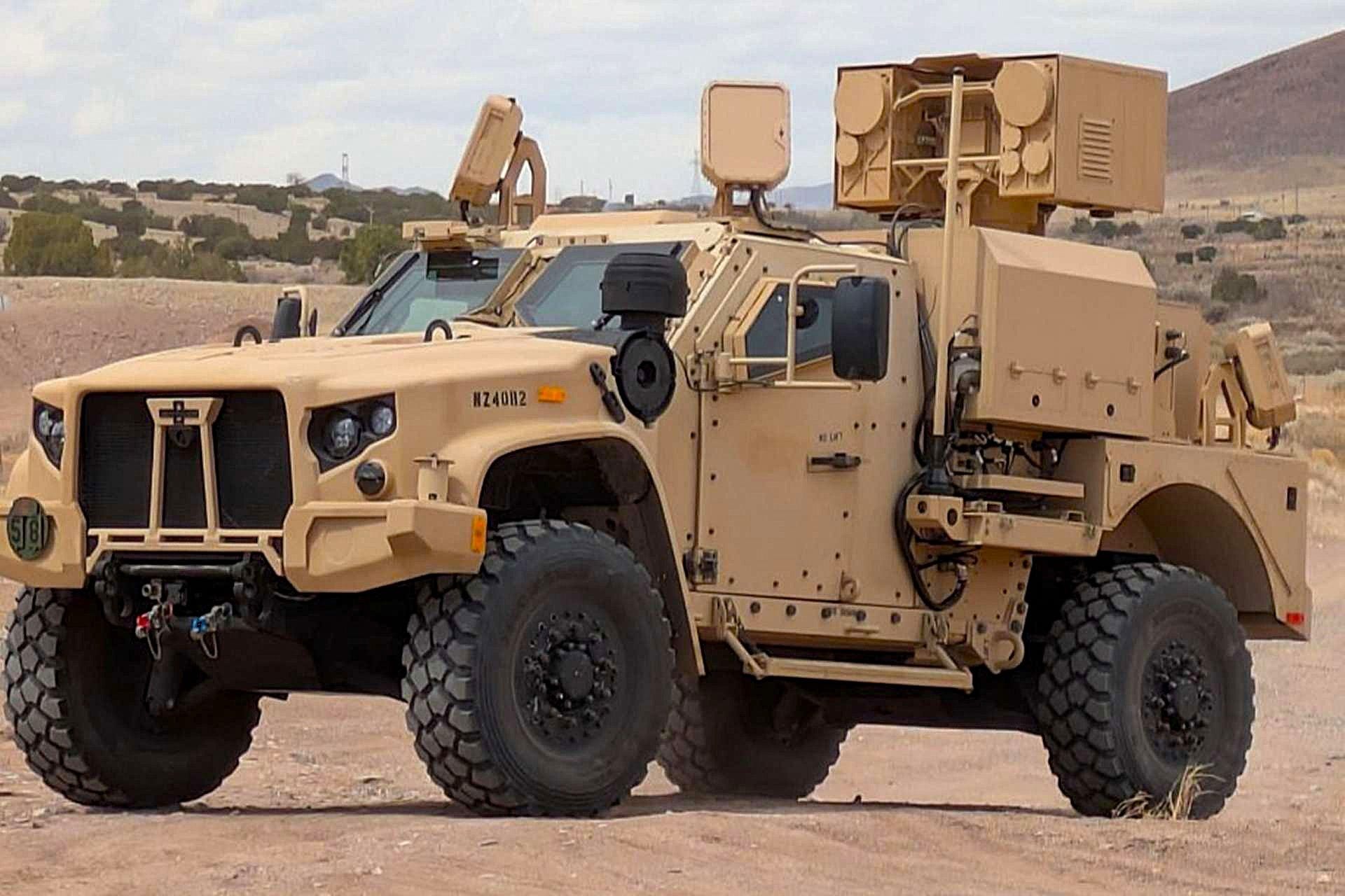 A US Army JLTV carries a LOCUST high-energy laser counter-drone system, its beam director and equipment raised on a rear mount, in a desert range.