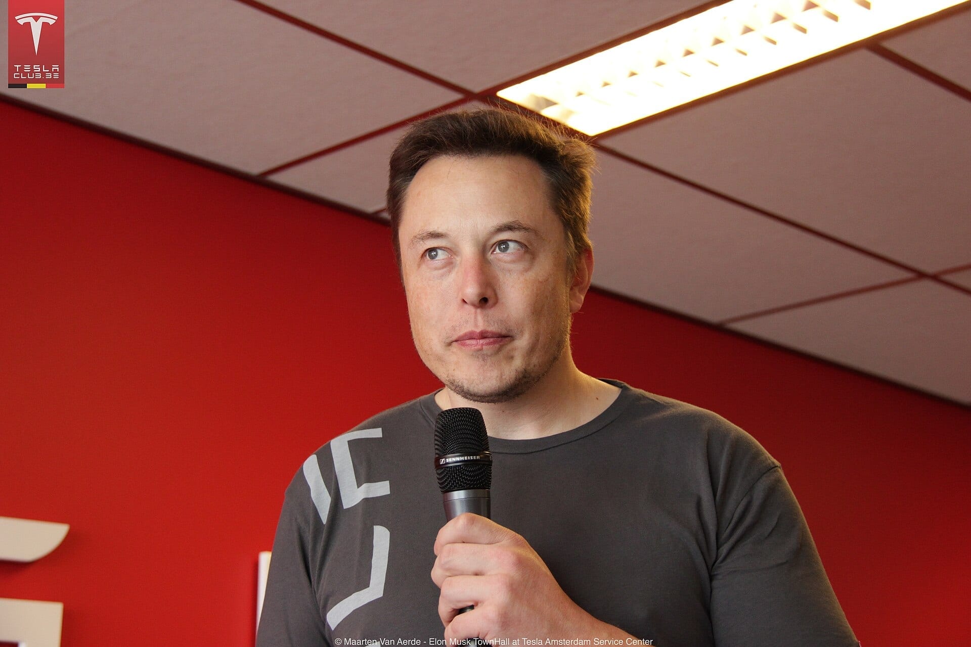 Elon Musk holding a microphone at a Tesla town hall event in Amsterdam, against a red wall.