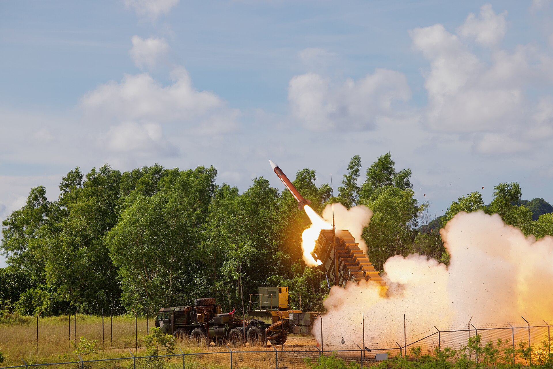 A Patriot interceptor missile firing from an M903 launching station in a grassy field, flames and smoke billowing around the canister.