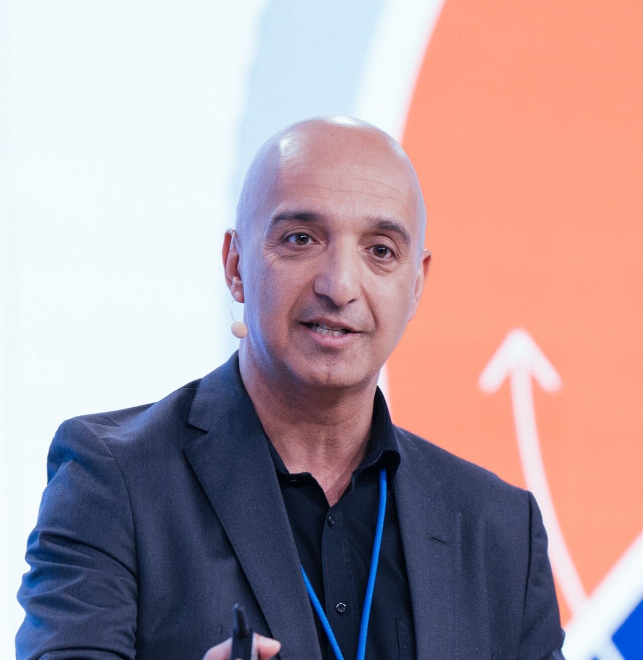 Novo Nordisk CEO Mike Doustdar speaking on a conference stage, wearing a headset microphone and dark blazer against an orange backdrop.