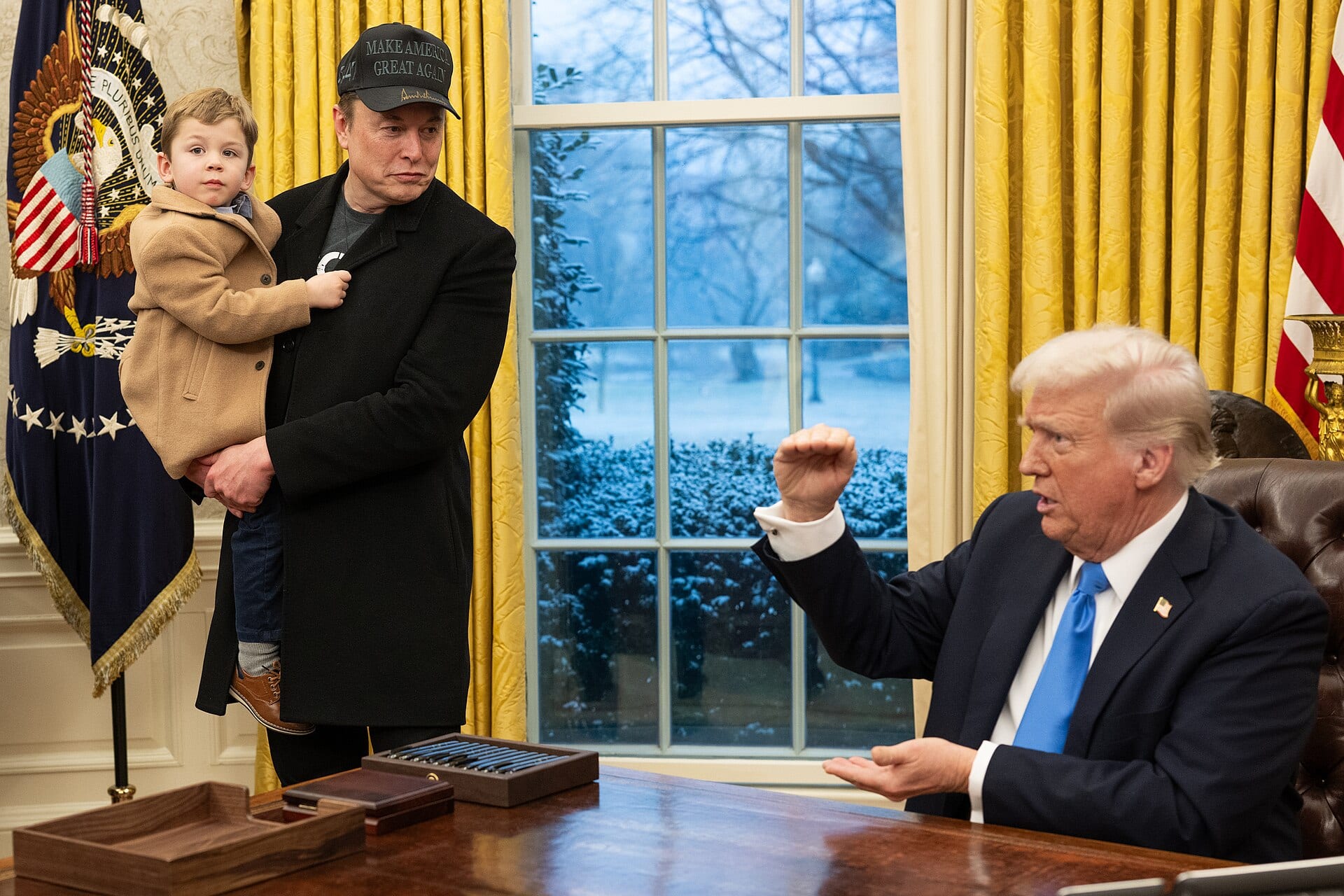 Elon Musk, in a MAGA cap and holding his son, watches Donald Trump sign executive orders in the Oval Office