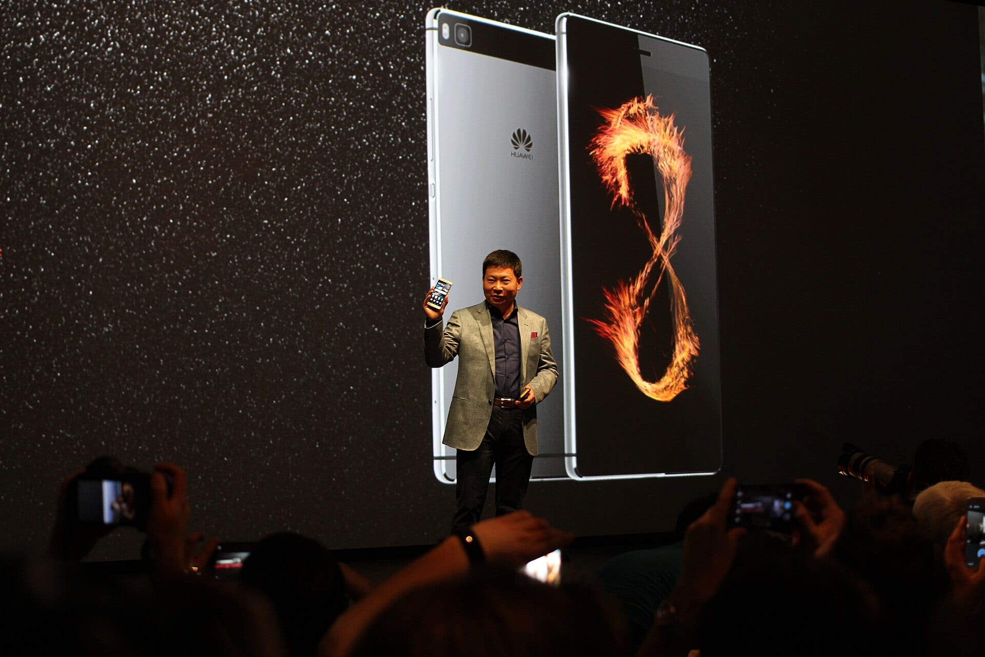 Richard Yu on a launch stage holding a Huawei smartphone, with giant images of the phone projected behind him