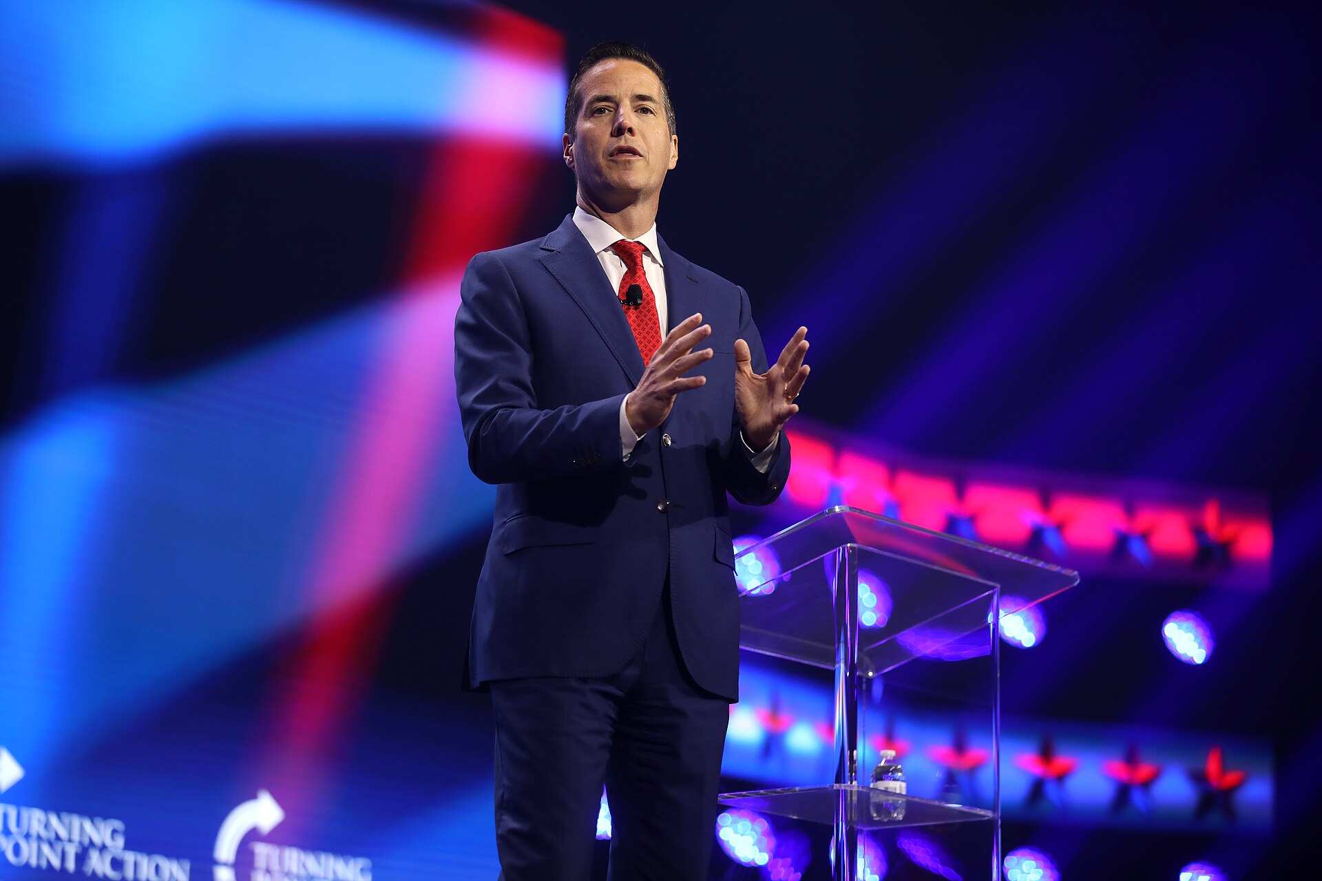 Bernie Moreno speaking on stage in a navy suit and red tie