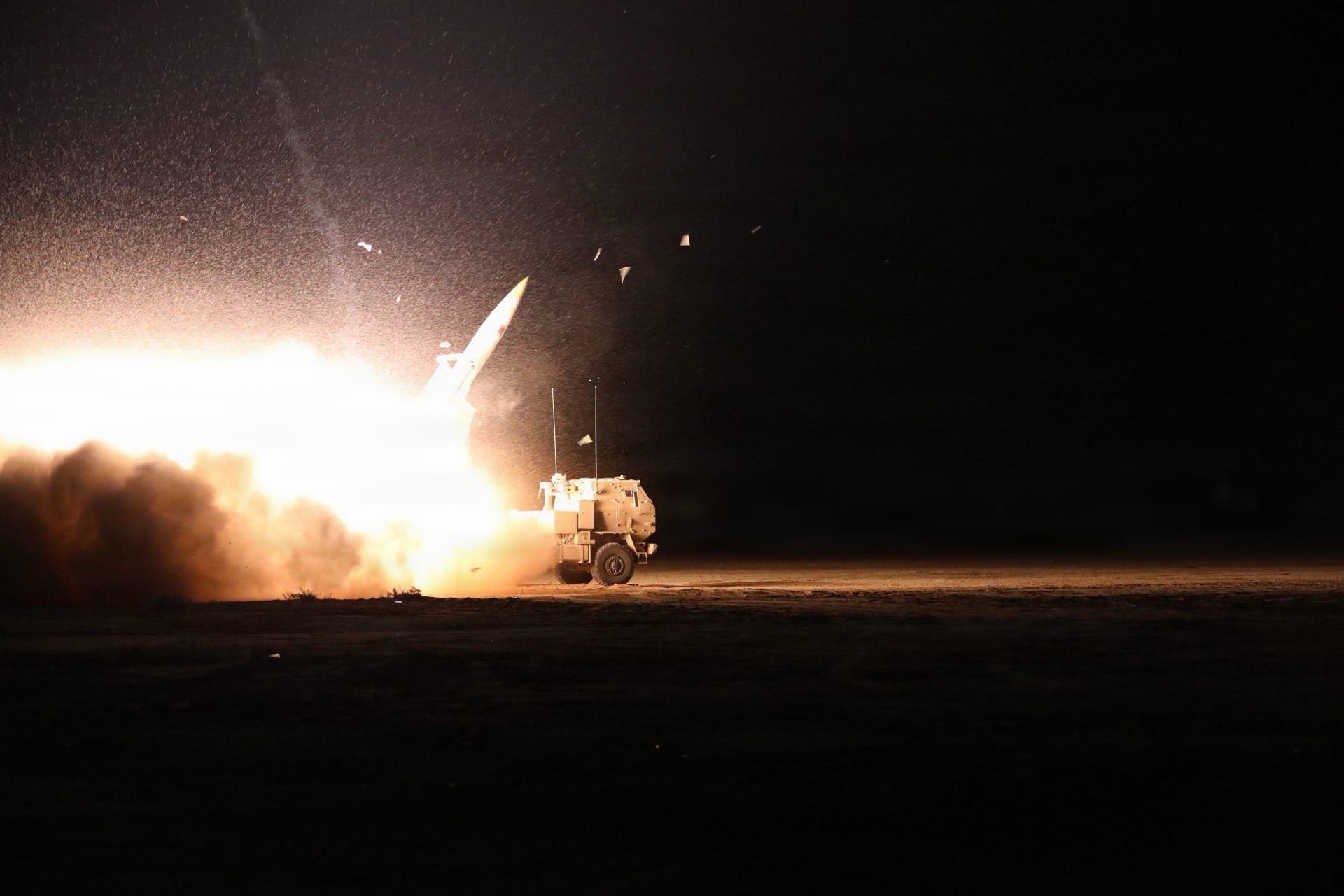 A HIMARS launcher fires a rocket at night, the launch flash lighting up the vehicle and the desert floor