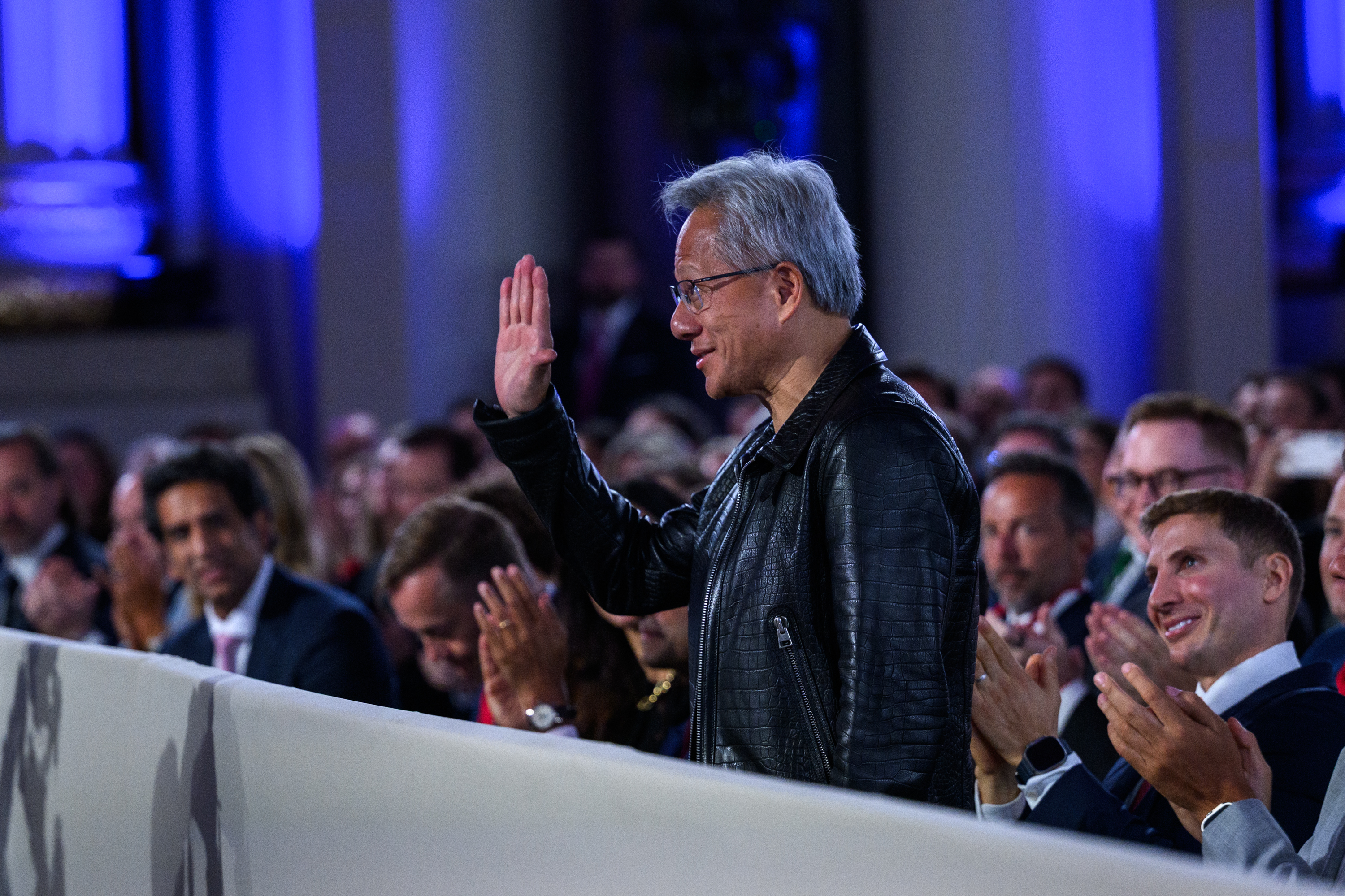 Nvidia CEO Jensen Huang, in a black leather jacket, stands and waves as he is recognized by President Trump during the White House AI Summit, while audience members applaud.