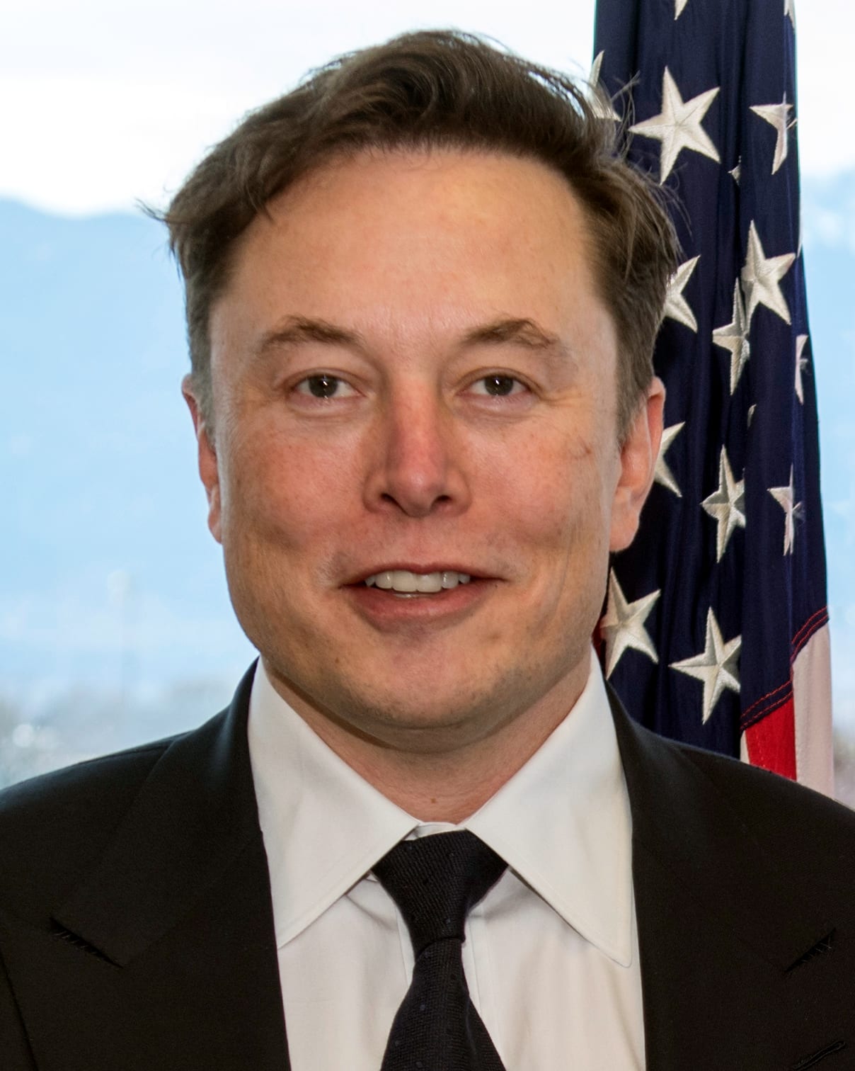 Elon Musk in a dark suit and tie, photographed in 2019 in front of a US flag