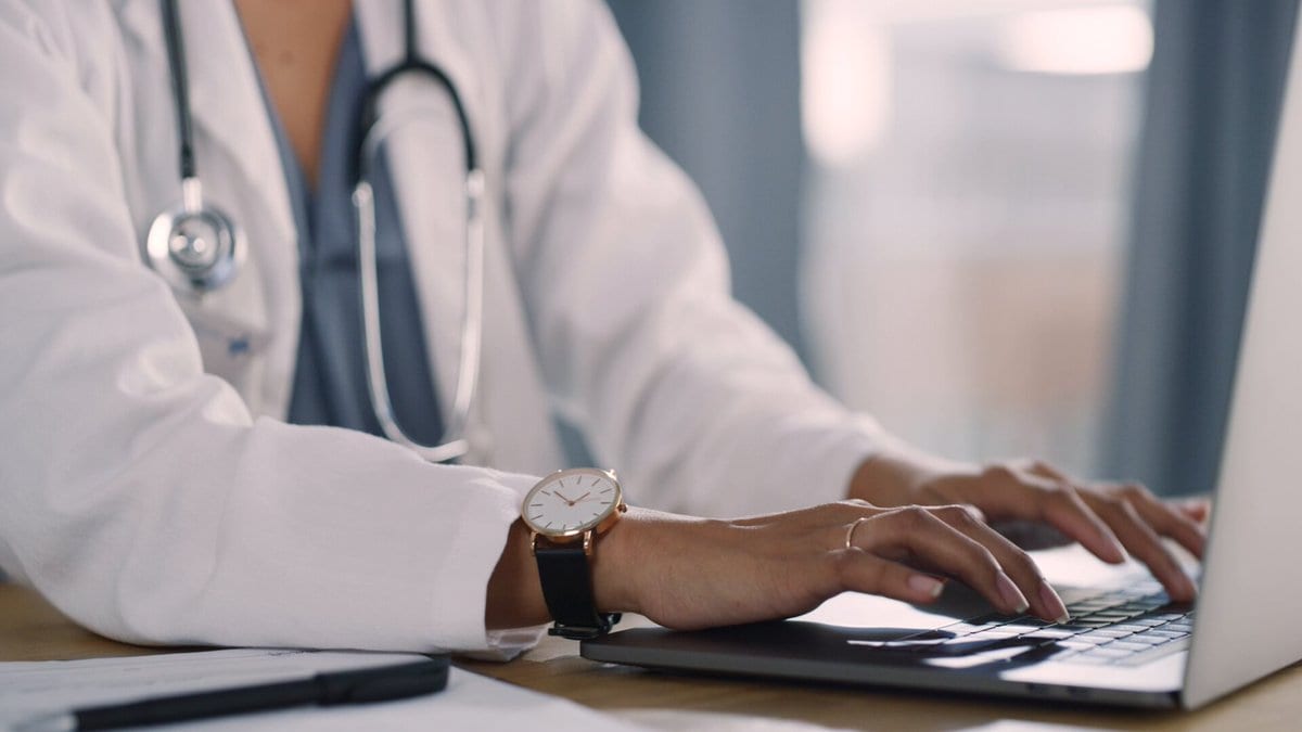 A physician in a white coat works at a laptop running Doximity's Clinical AI Suite