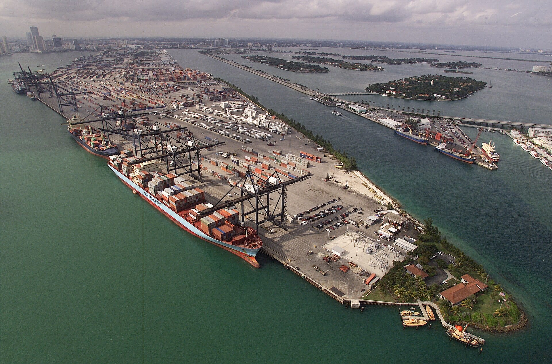 Aerial view of a container port with gantry cranes, stacked shipping containers and a loaded Maersk cargo ship at the dock.