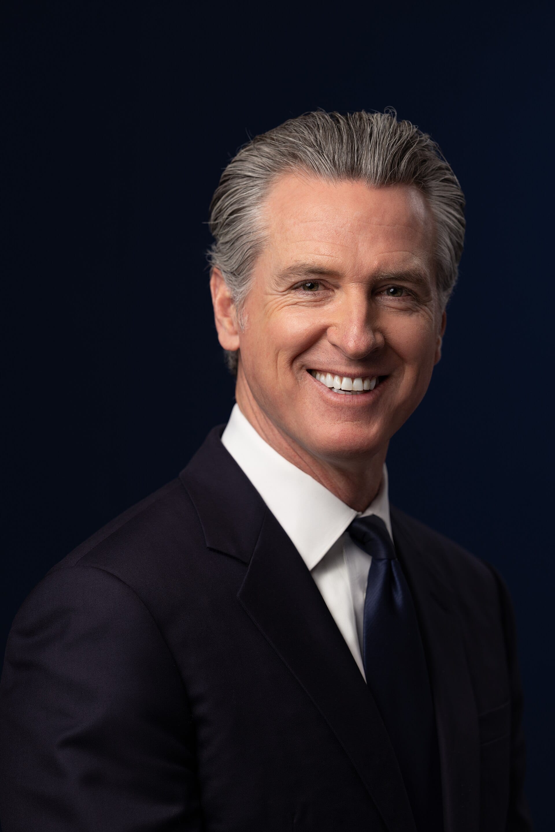 Gouverneur Gavin Newsom im dunklen Anzug und mit navyblauer Krawatte vor dunkelblauem Hintergrund, offizielles Porträt von 2026
