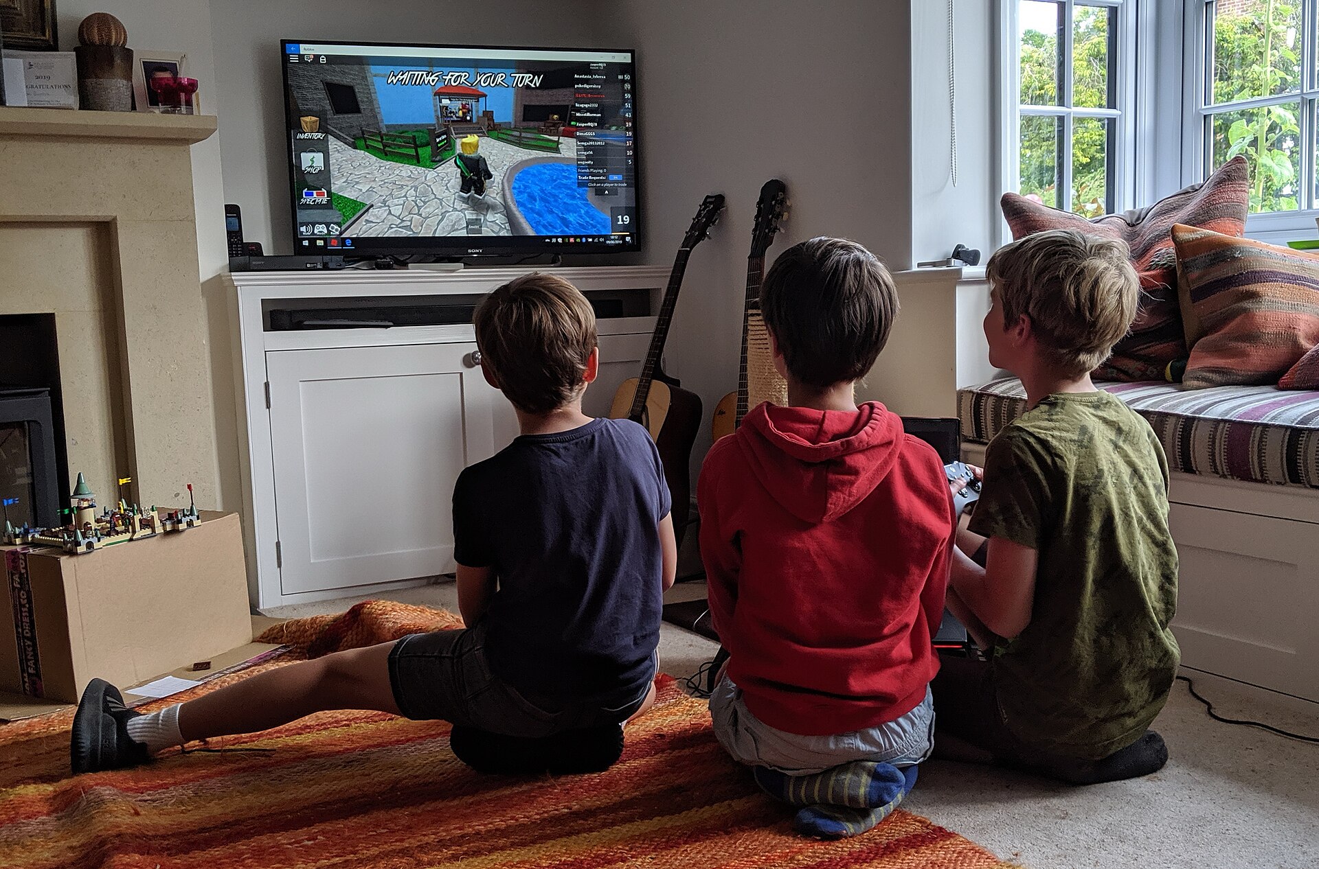 Three children playing Roblox on a television in a living room, with a public server player list visible on screen
