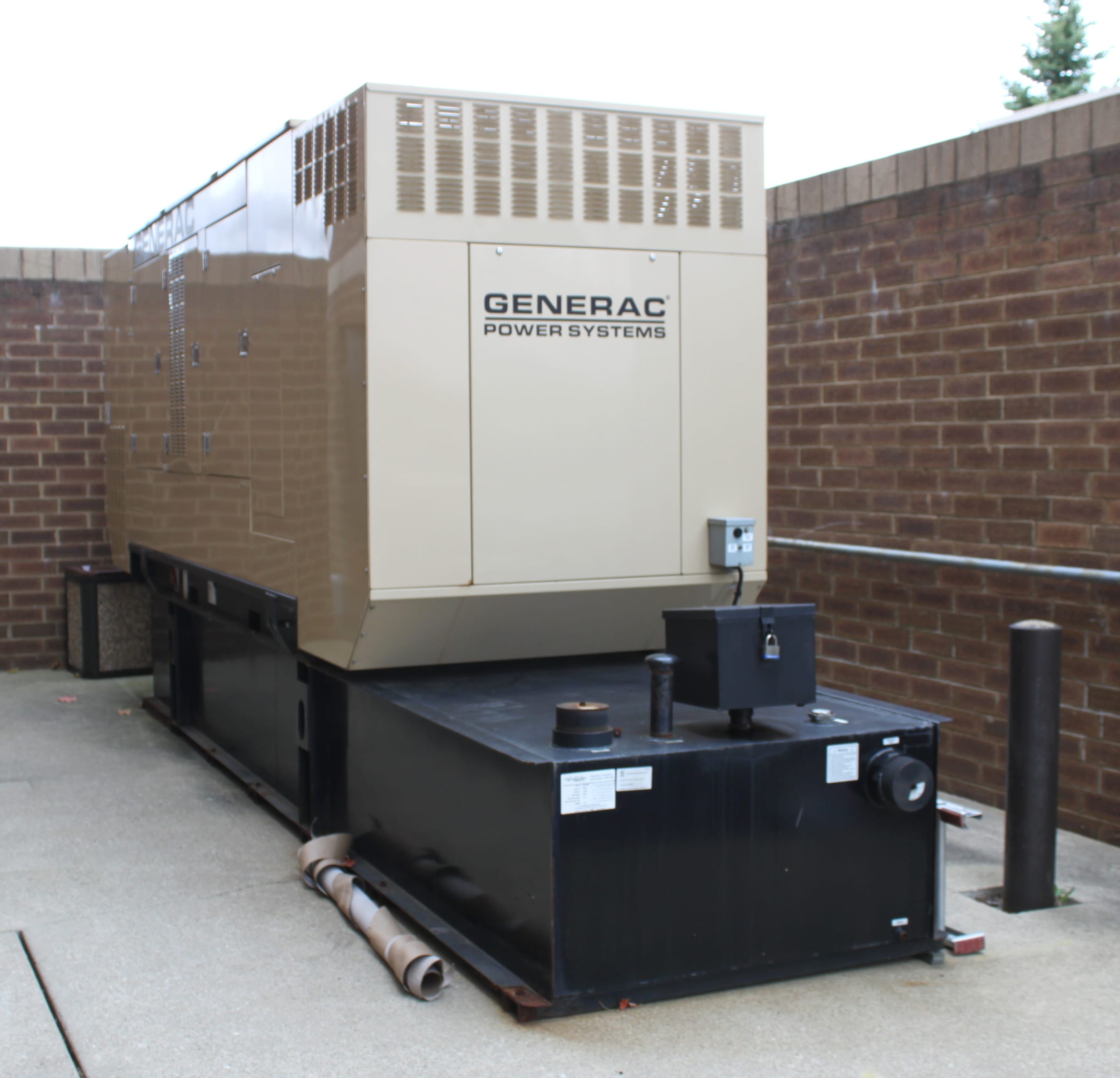 A large beige Generac Power Systems commercial backup generator installed outdoors against a brick wall