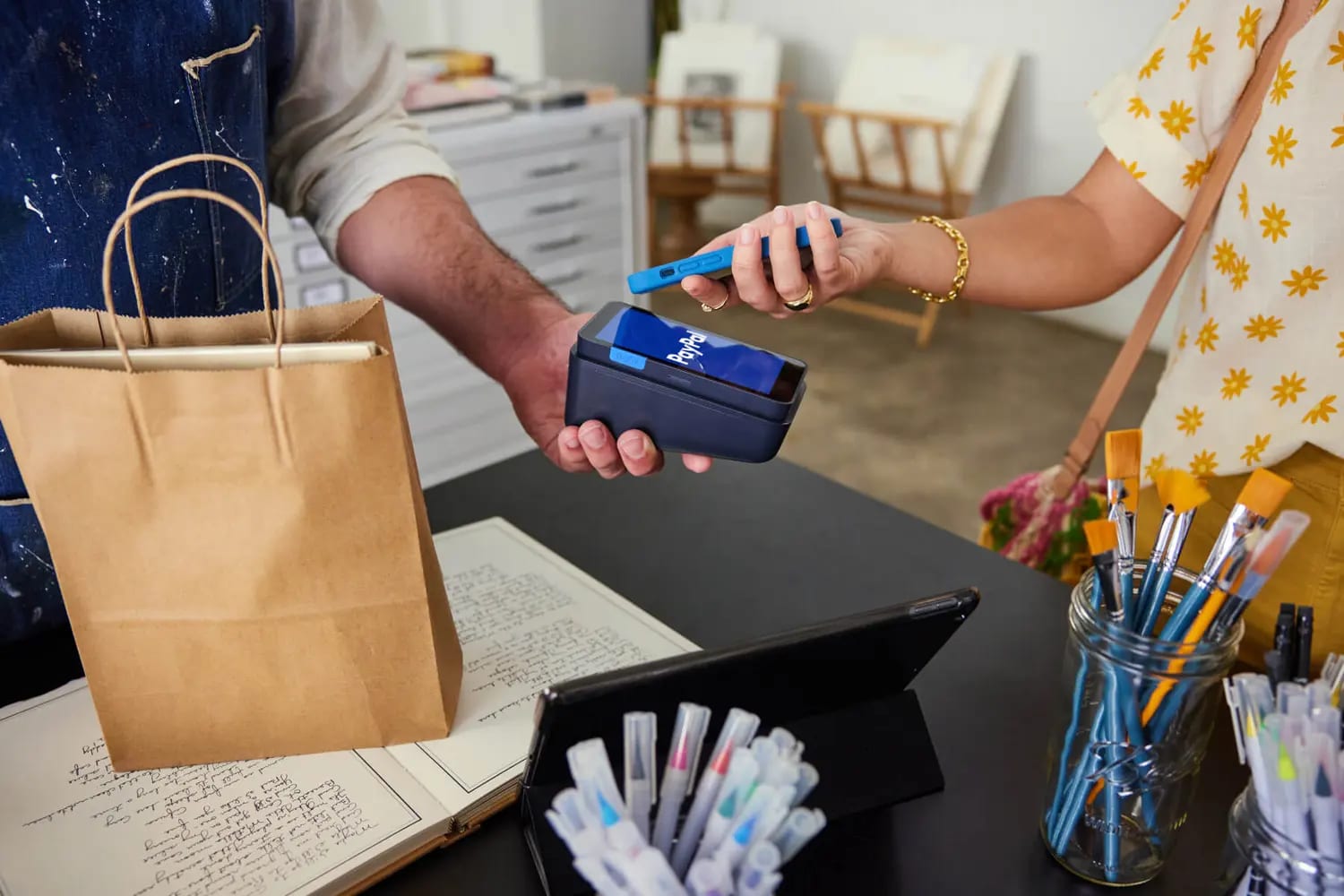A customer pays by tapping a phone on a PayPal-branded card reader at a shop counter