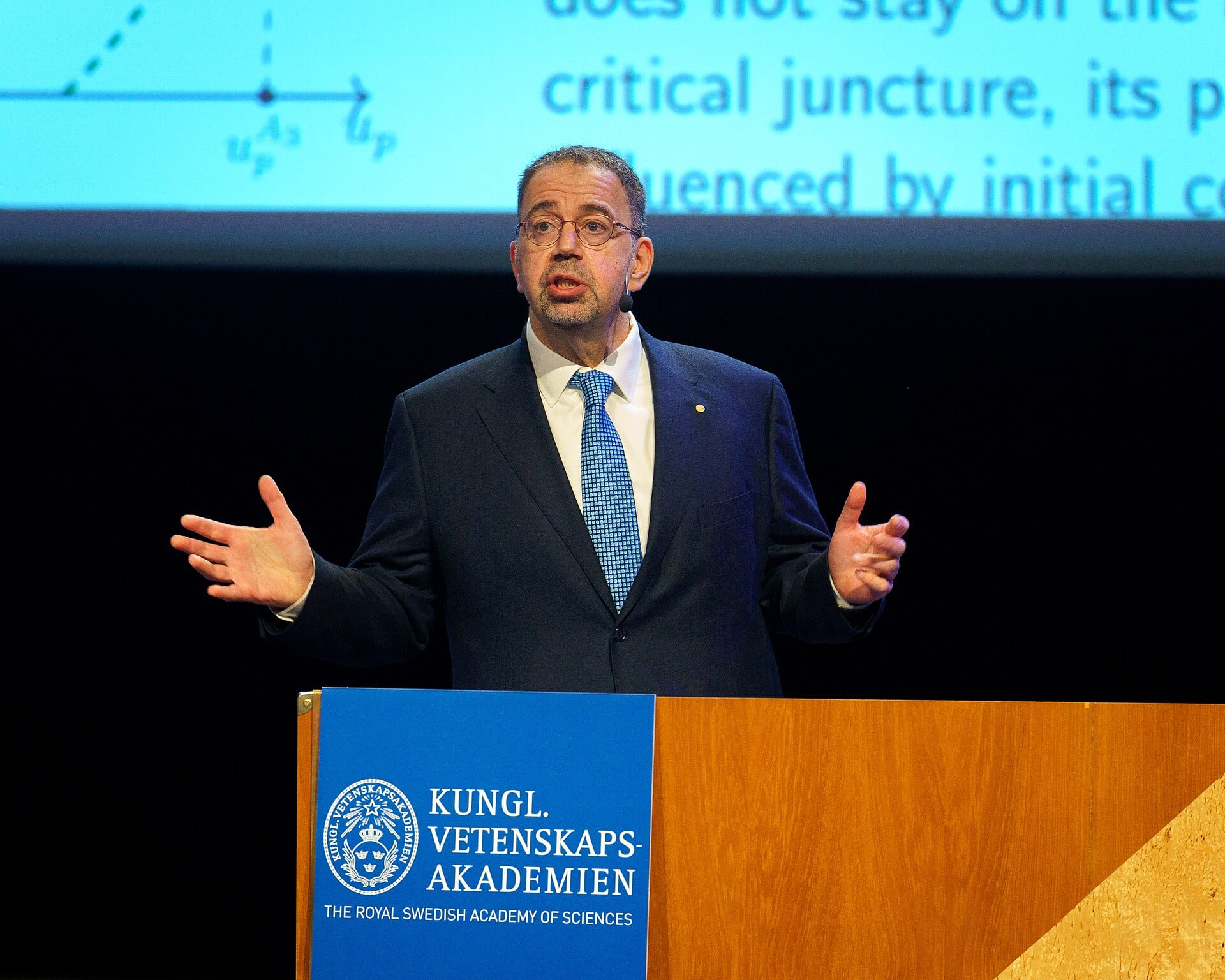 Daron Acemoglu speaking at a lectern at the Royal Swedish Academy of Sciences during his Nobel lectures