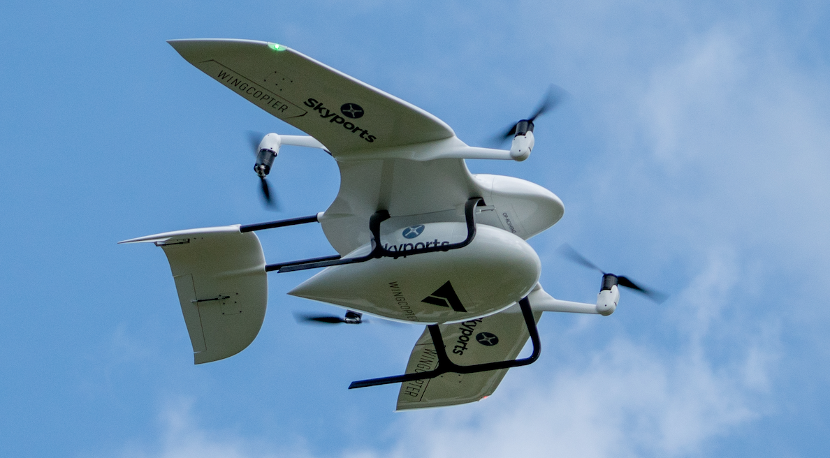 A Skyports-branded Wingcopter cargo drone banking in flight against a blue sky