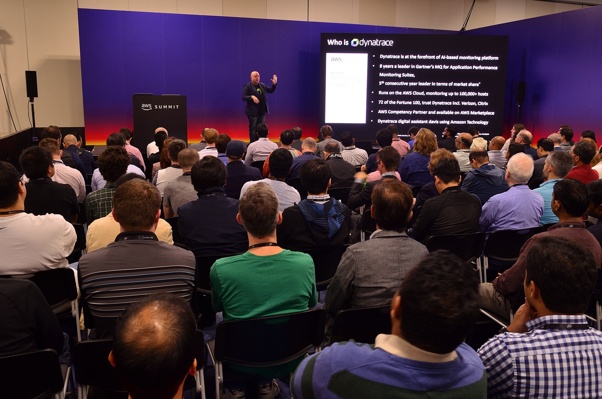 A presenter on an AWS Summit stage beside a slide titled Who is Dynatrace, in front of a seated audience