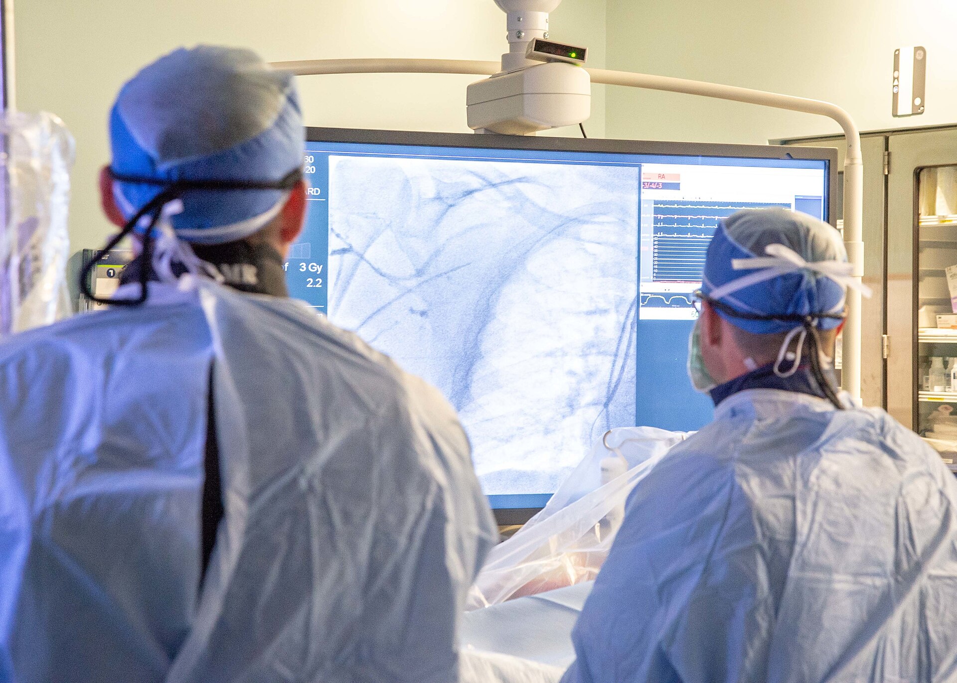 Clinicians in surgical caps watch live fluoroscopic coronary images on a large display during a cardiac catheterization procedure.