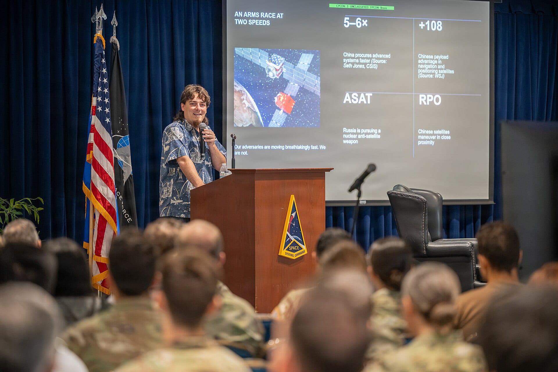 Palmer Luckey speaking into a microphone at a podium before a Space Systems Command audience, with a slide titled An Arms Race at Two Speeds behind him