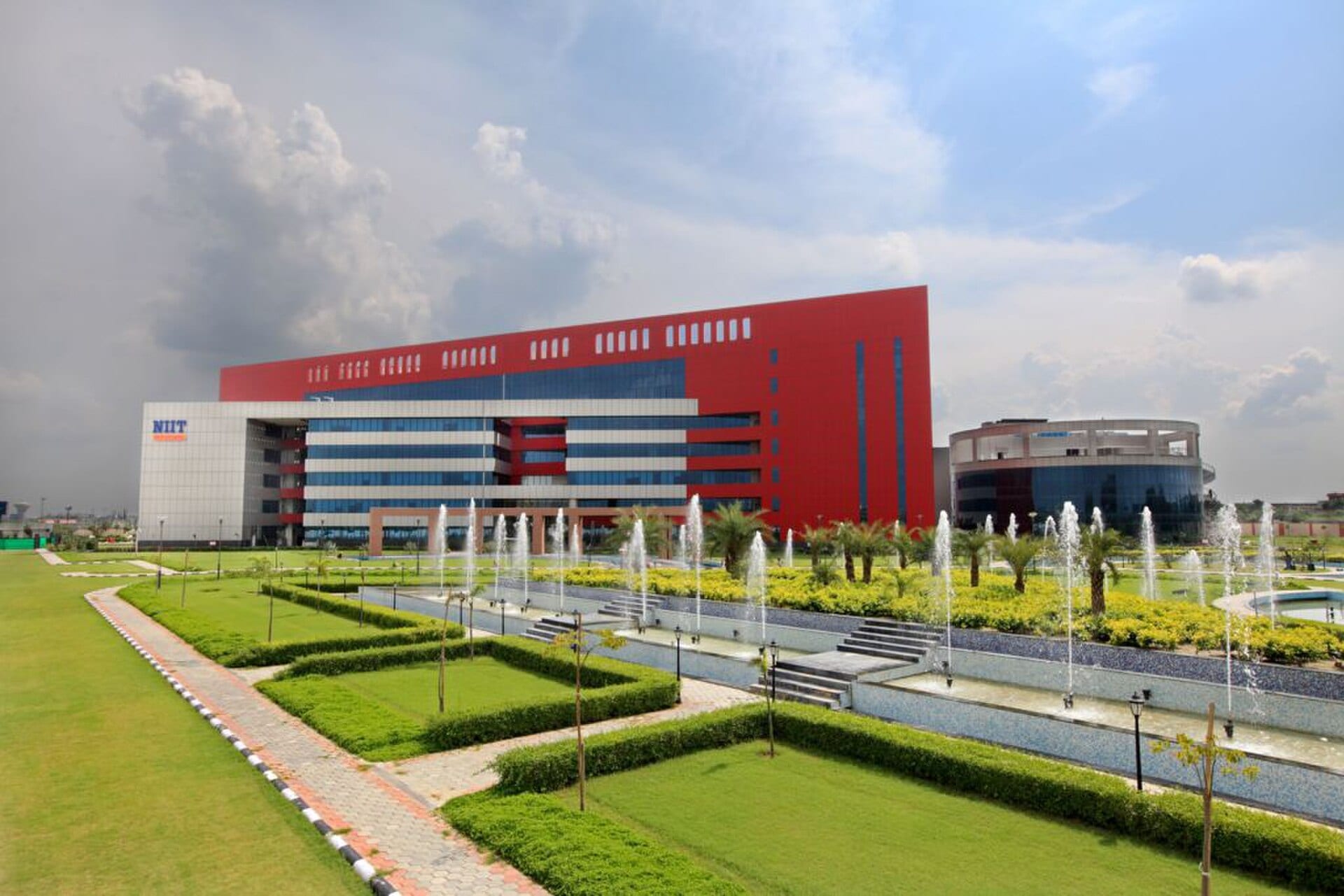The red and white office tower of the NIIT Technologies campus at Greater Noida, with lawns, hedges and fountains in front
