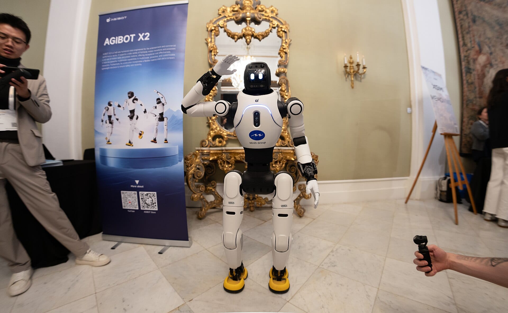 An AgiBot X2 humanoid robot waves at an exhibition stand bearing its name