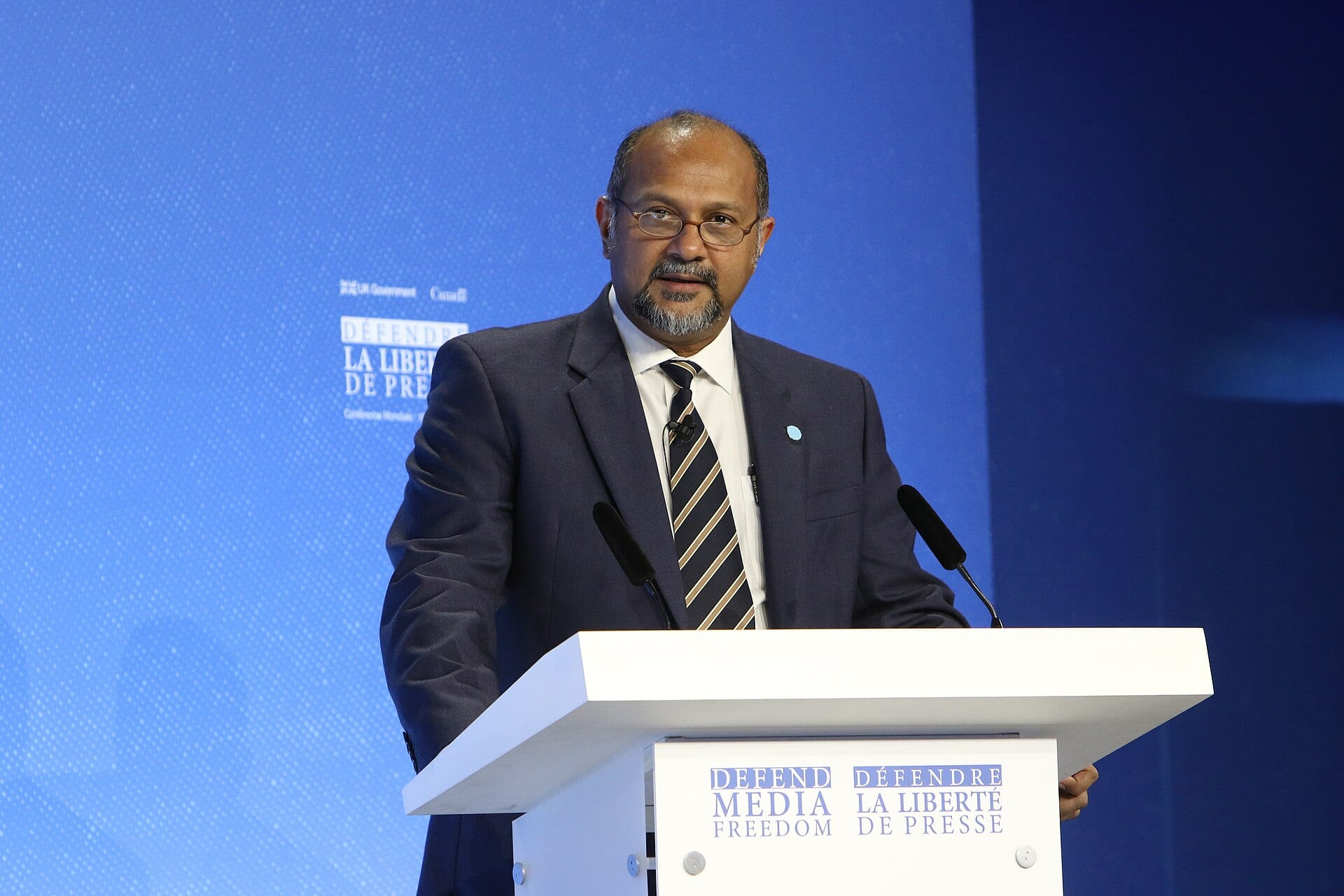 Gobind Singh Deo speaking at a conference podium in a dark suit and striped tie