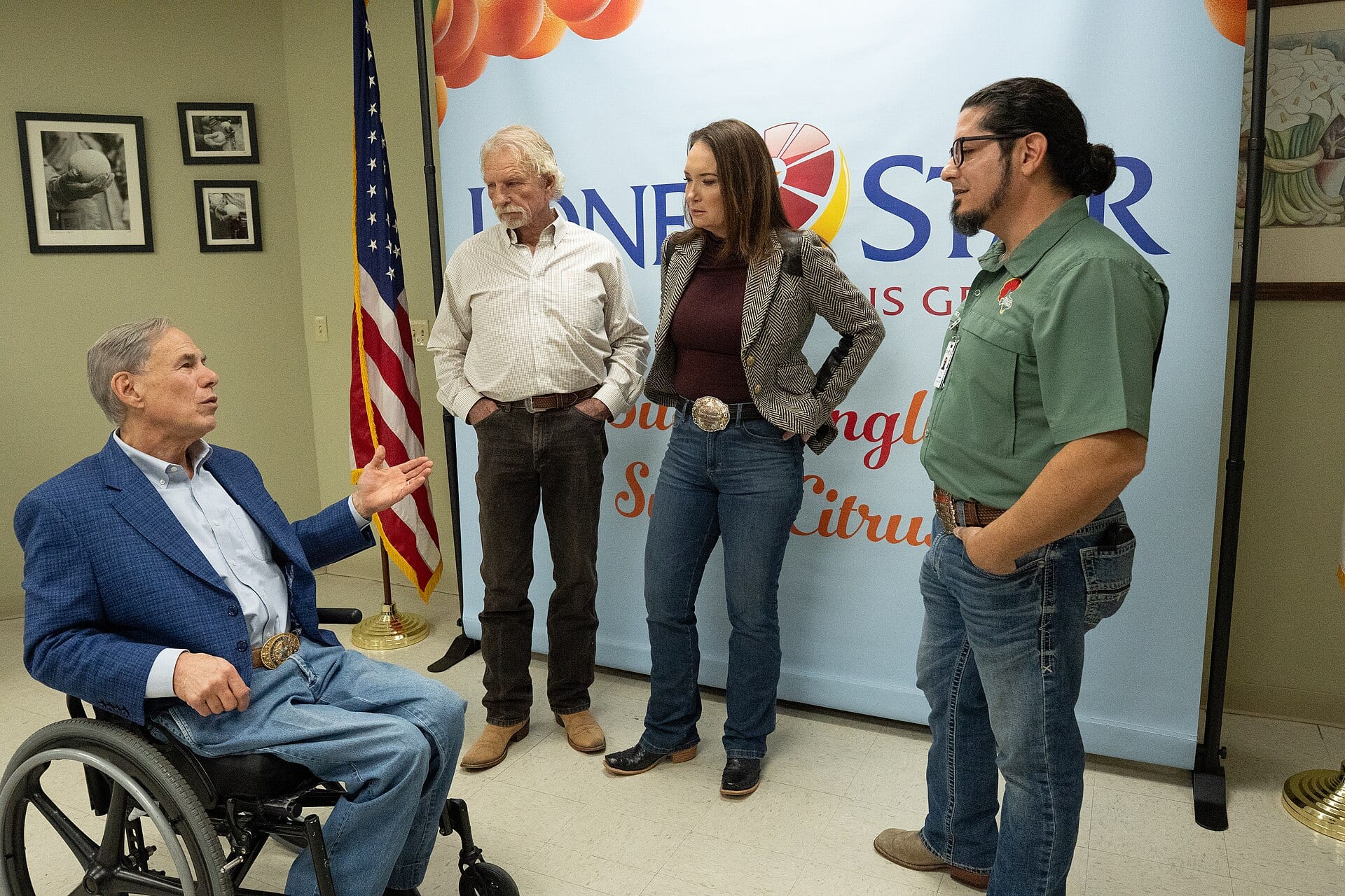 Texas Governor Greg Abbott, seated in a blue blazer and jeans, gestures while speaking with growers and officials in front of a Lone Star Citrus Growers banner and an American flag.