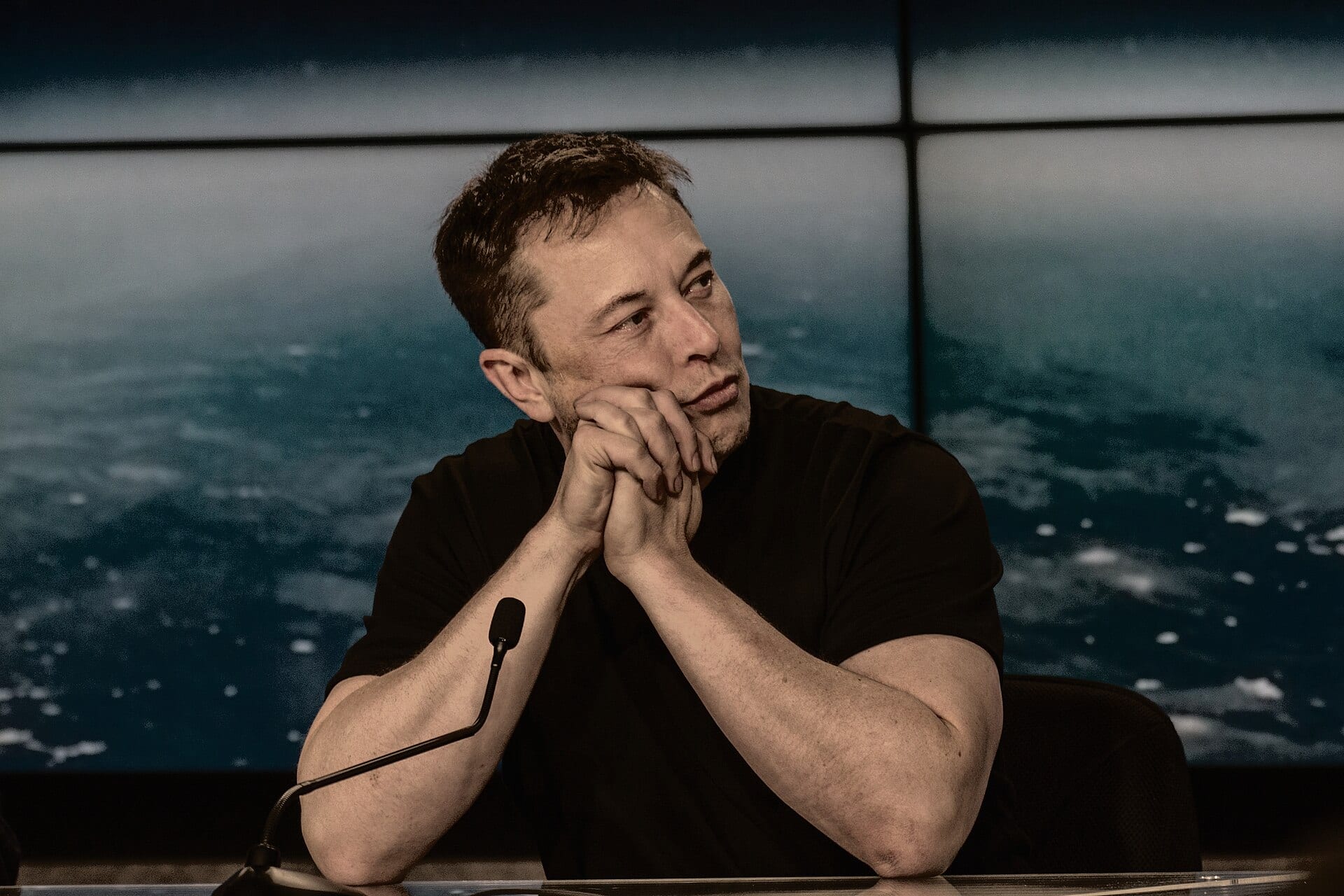 Elon Musk listening at a press conference, an image of Earth from space on the screen behind him