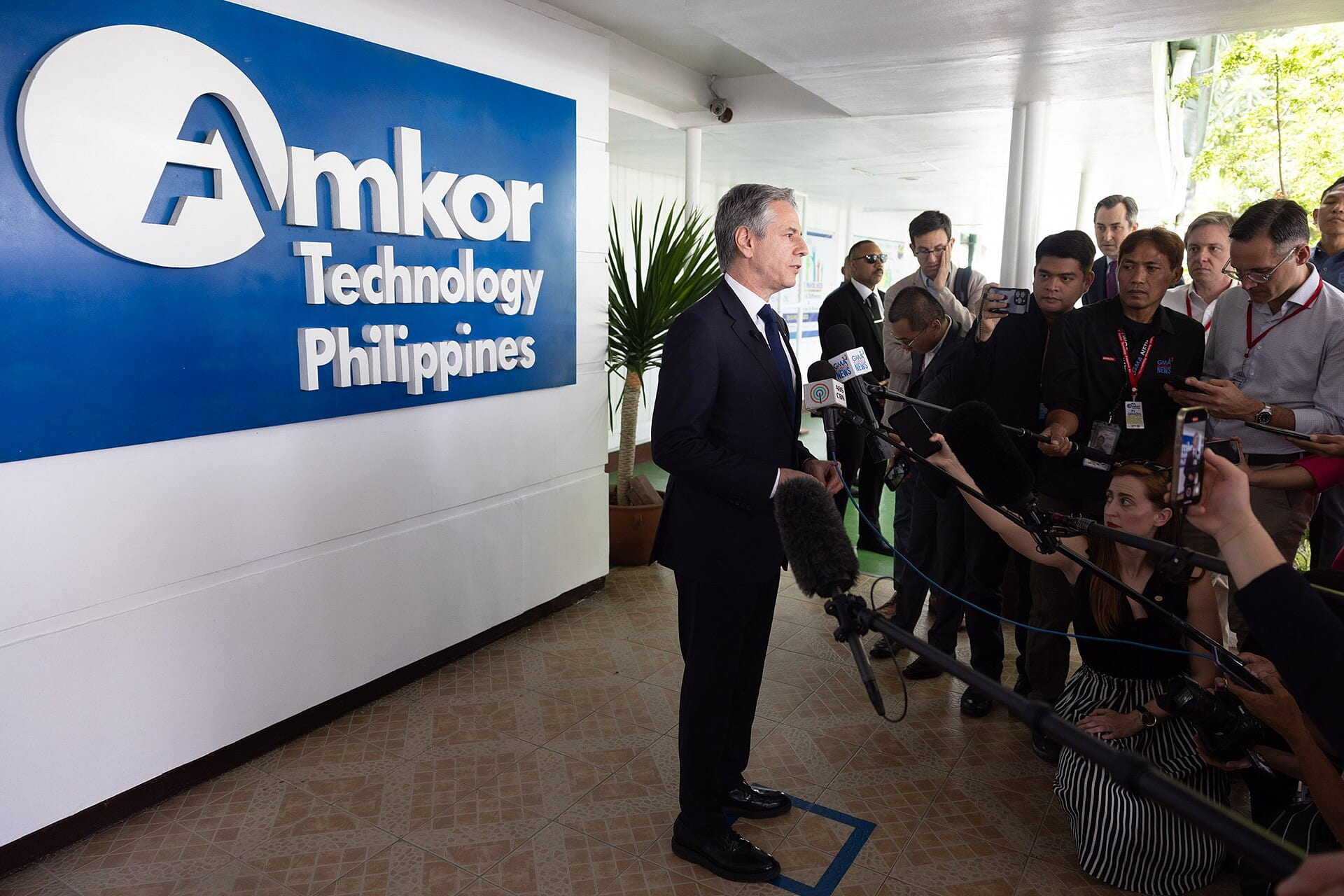 U.S. Secretary of State Antony Blinken faces reporters and microphones in front of a large blue Amkor Technology Philippines sign at the company's plant.