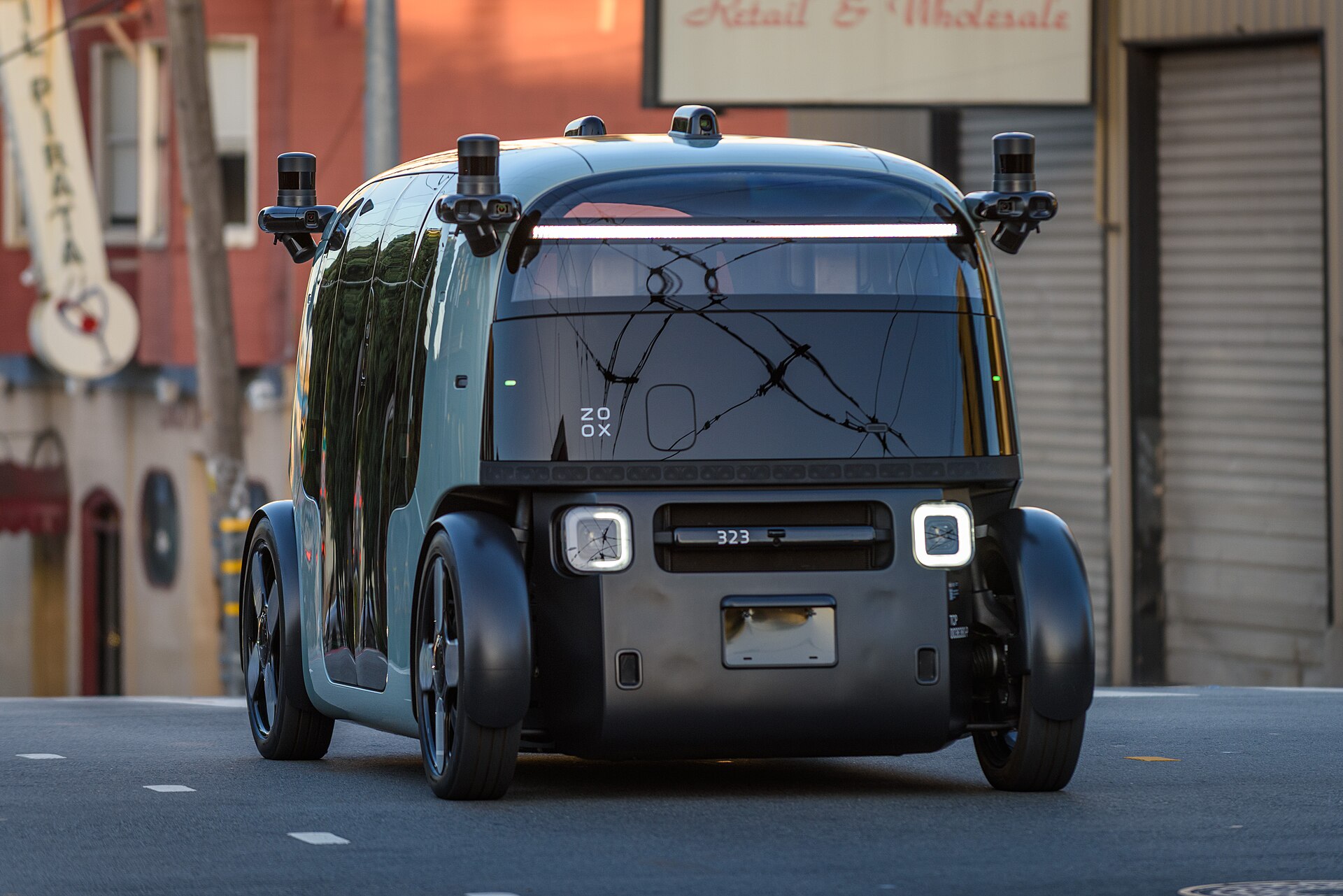 A dark green purpose-built Zoox robotaxi with corner-mounted sensors and no steering wheel drives on a San Francisco street.