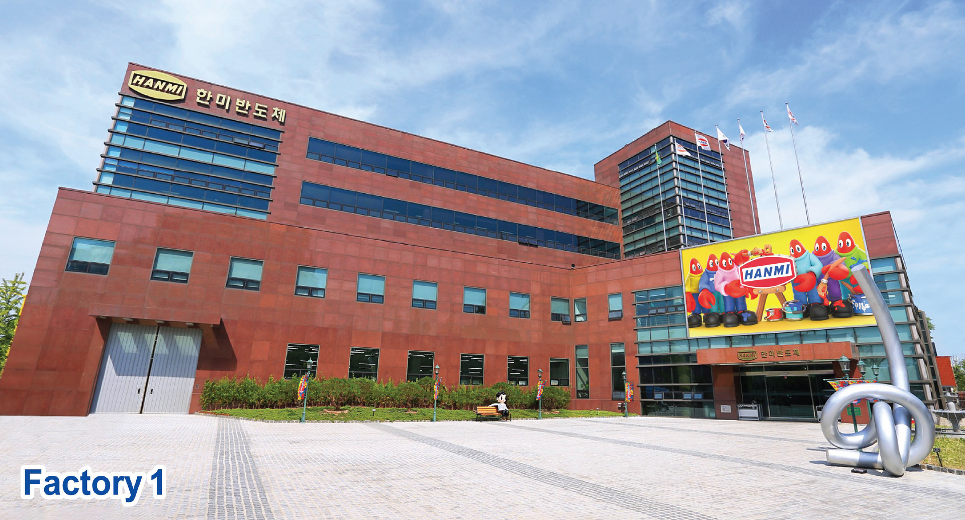 Hanmi Semiconductor's Factory 1 building, a red-brown plant with Hanmi signage, flags and a company mascot billboard
