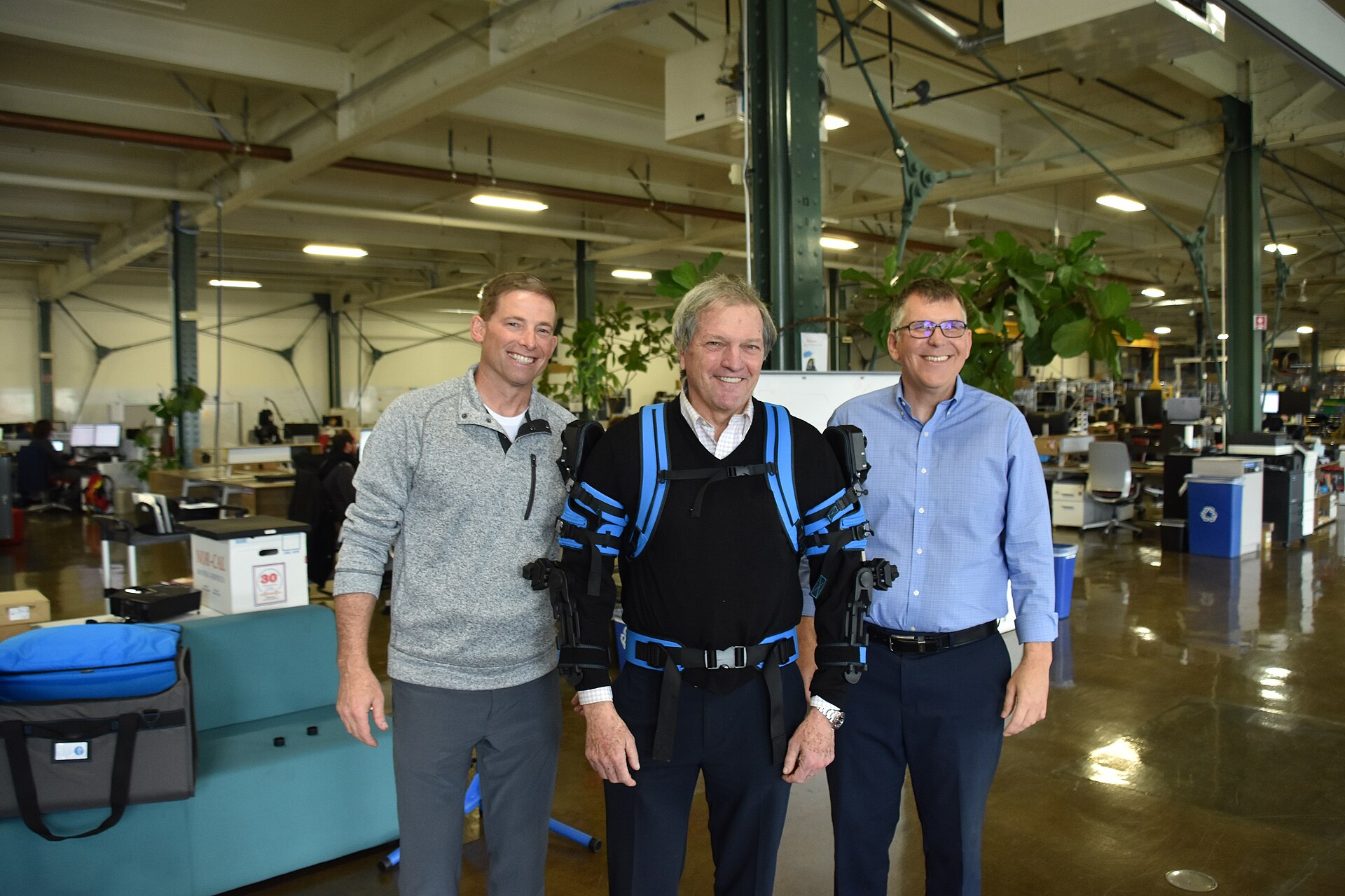 A man wearing an Ekso Bionics upper-body exoskeleton, flanked by two other men in an industrial workshop