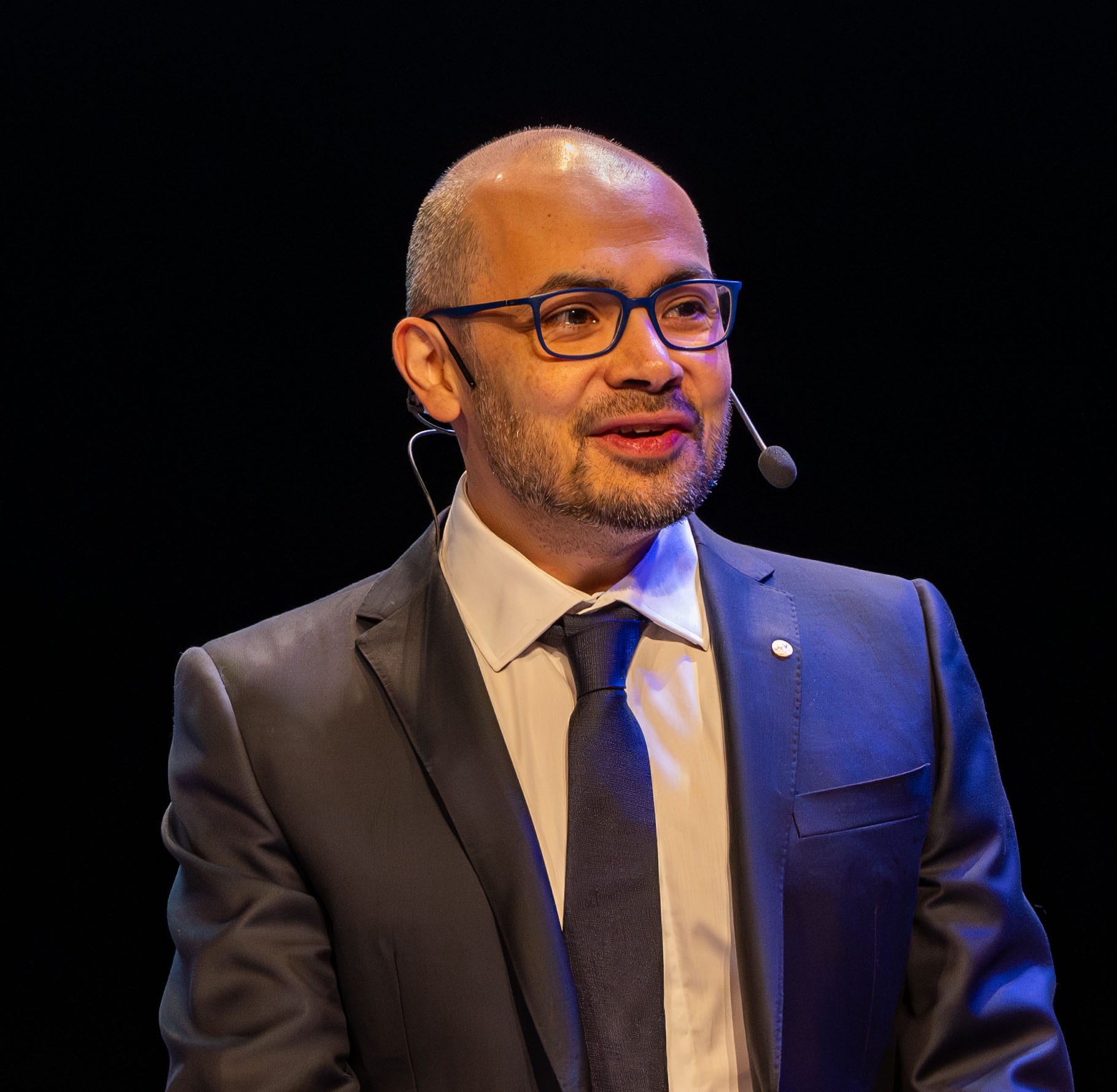 Demis Hassabis, cofounder of Google DeepMind, speaking at the 2024 Nobel Prize events in a dark suit and headset microphone against a black background.