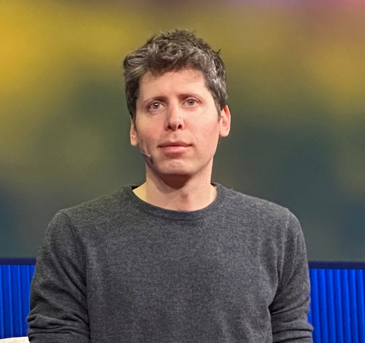 Sam Altman seated on stage during a TED talk, wearing a grey sweater and looking off to the side