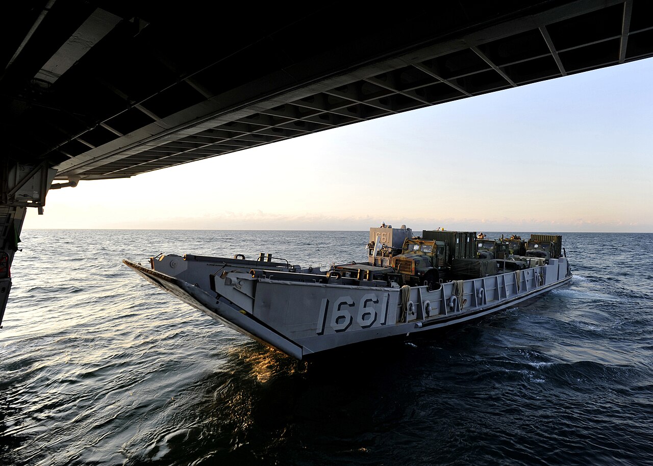 A gray Landing Craft Utility numbered 1661 carries vehicles as it approaches the lowered stern gate of an amphibious warship at sunset.
