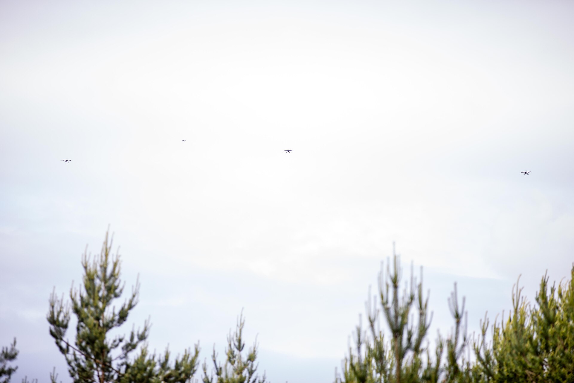Small drones fly in an overcast sky above treetops