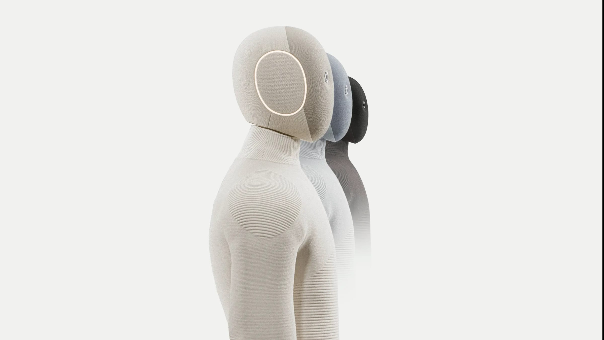 Three 1X NEO home robots in tan, blue and black stand in a row against a pale background