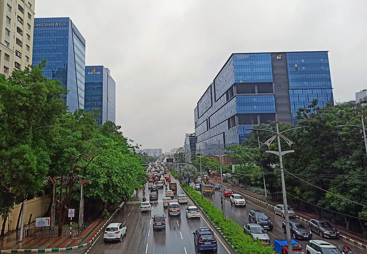 Blue-glass office towers over a busy Hyderabad corridor, one carrying the Sattva brand and another JPMorgan Chase signage