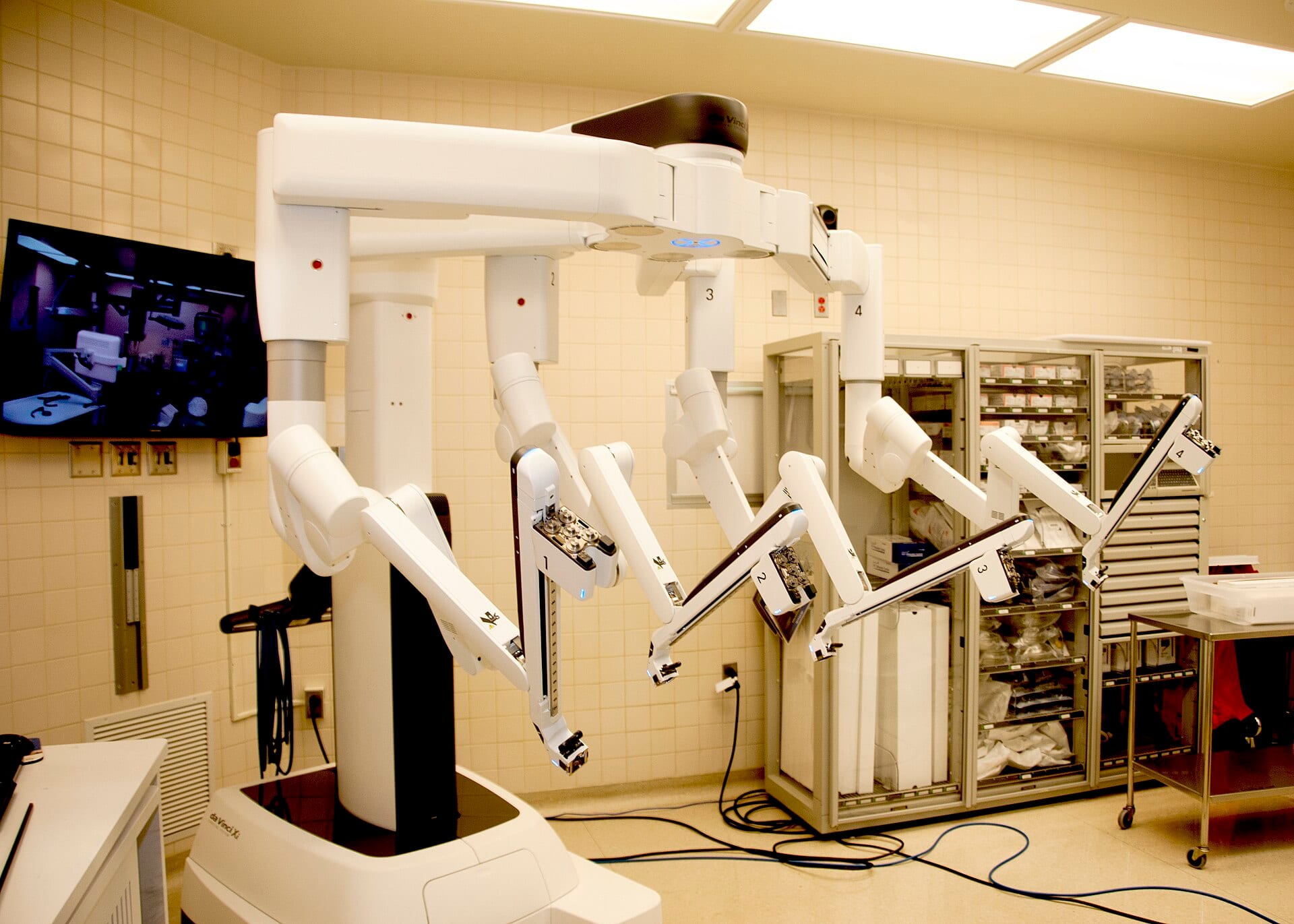 A da Vinci Xi surgical system with its four instrument arms mounted stands ready in an operating room, beside a cabinet of wrapped surgical instruments.