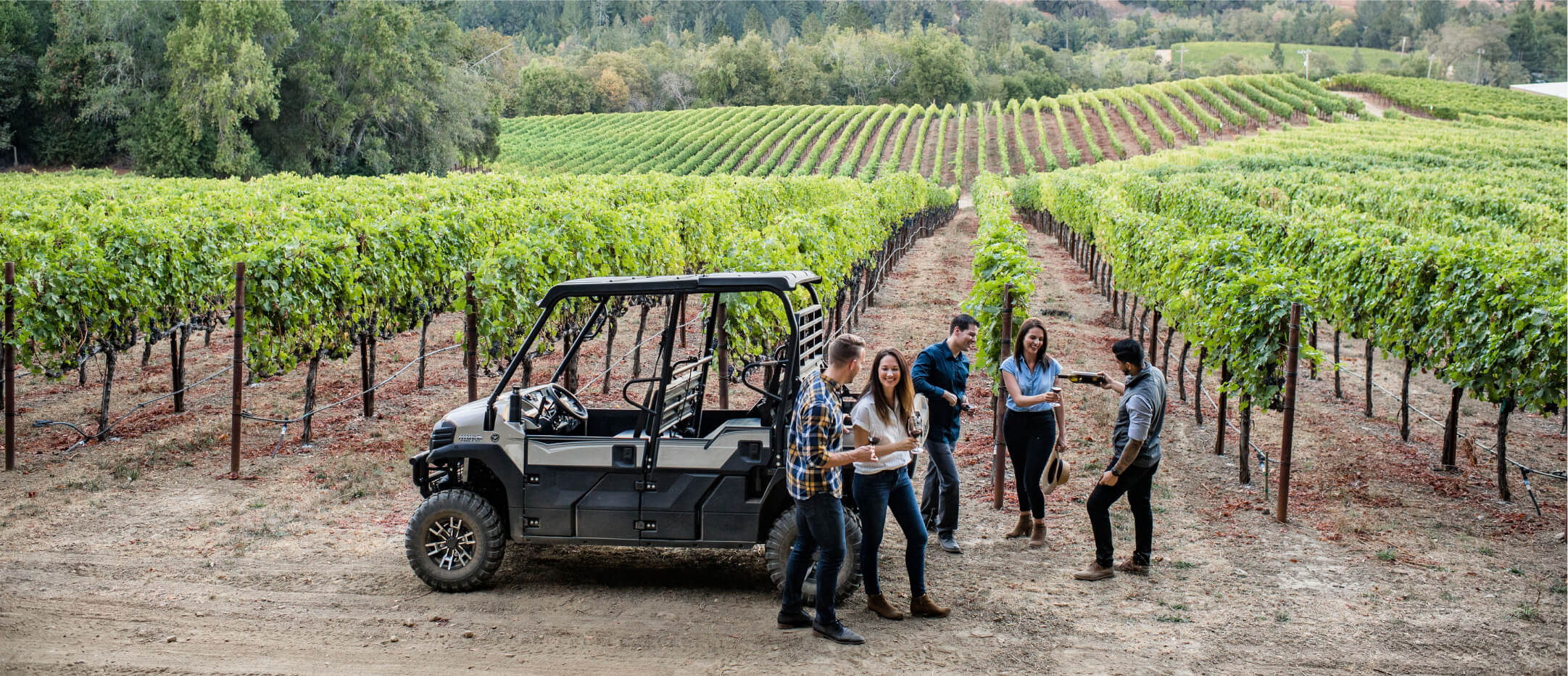 Wide view of five individuals standing and enjoying wine together on a vinyard next to a cross-terrain vehicle.
