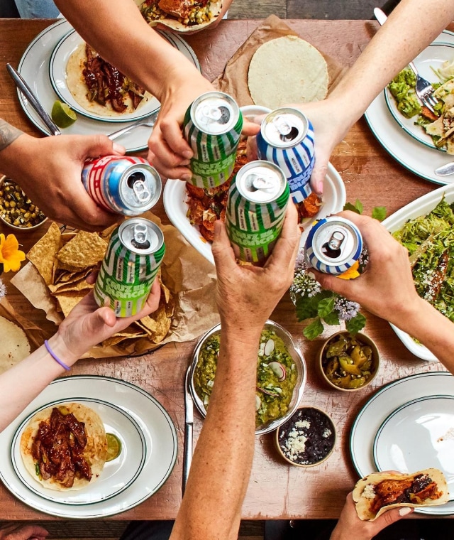 Overhead shot of six arms cheering canned drinks over a table covered with plates of tacos, chips, and toppings.