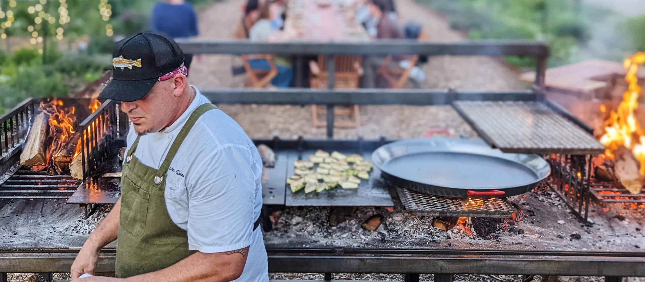 A chef cooking on an open fire with a table of people in the background.