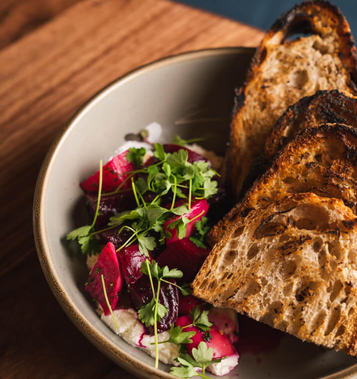 Four pieces of toast sit in a grey bowl. To the left of the toast is a white puree with a pile of bright red beets on top of it that is garnished with green veggies. Everything is on top of a wood-grained table.