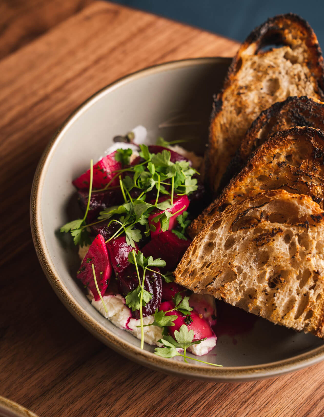 Four pieces of toast sit in a grey bowl. To the left of the toast is a white puree with a pile of bright red beets on top of it that is garnished with green veggies. Everything is on top of a wood-grained table.